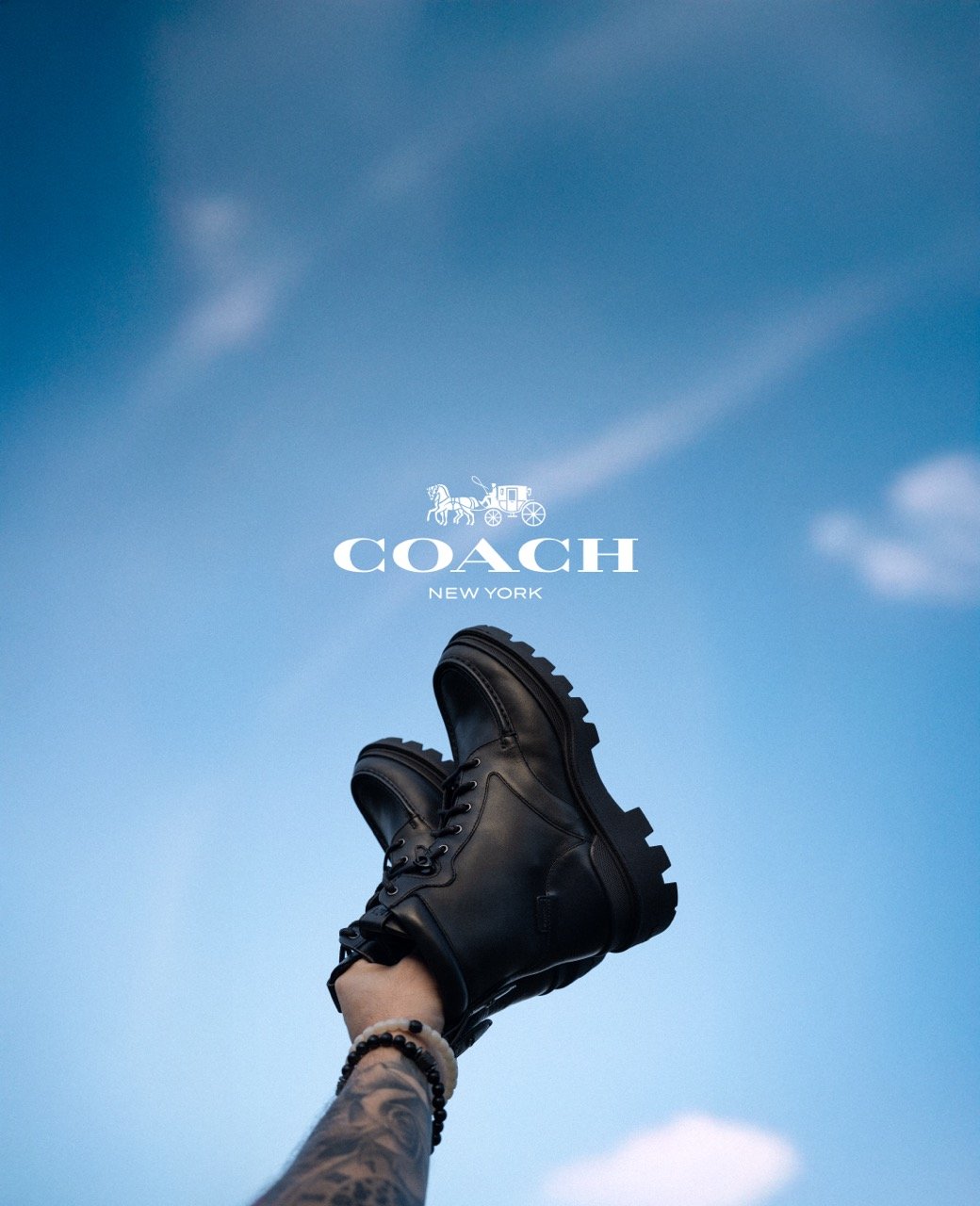 A person wearing black leather boots with a tattooed arm and beaded bracelet, holding the boots against a blue sky with clouds and the Coach New York logo.
