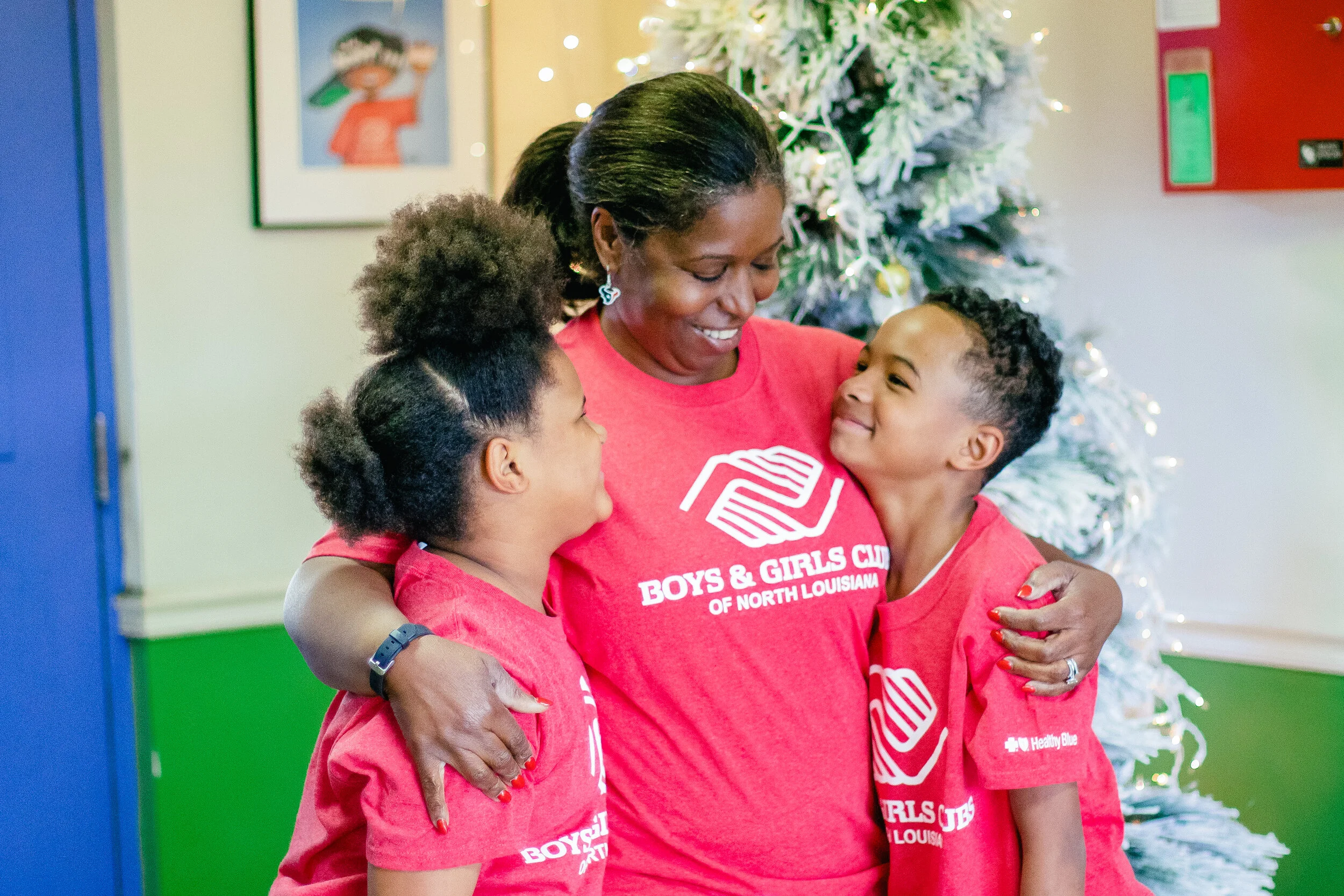An adult woman hugging two children, all wearing red shirts that say 'Boys & Girls Club of North Louisiana' with a Christmas tree in the background.