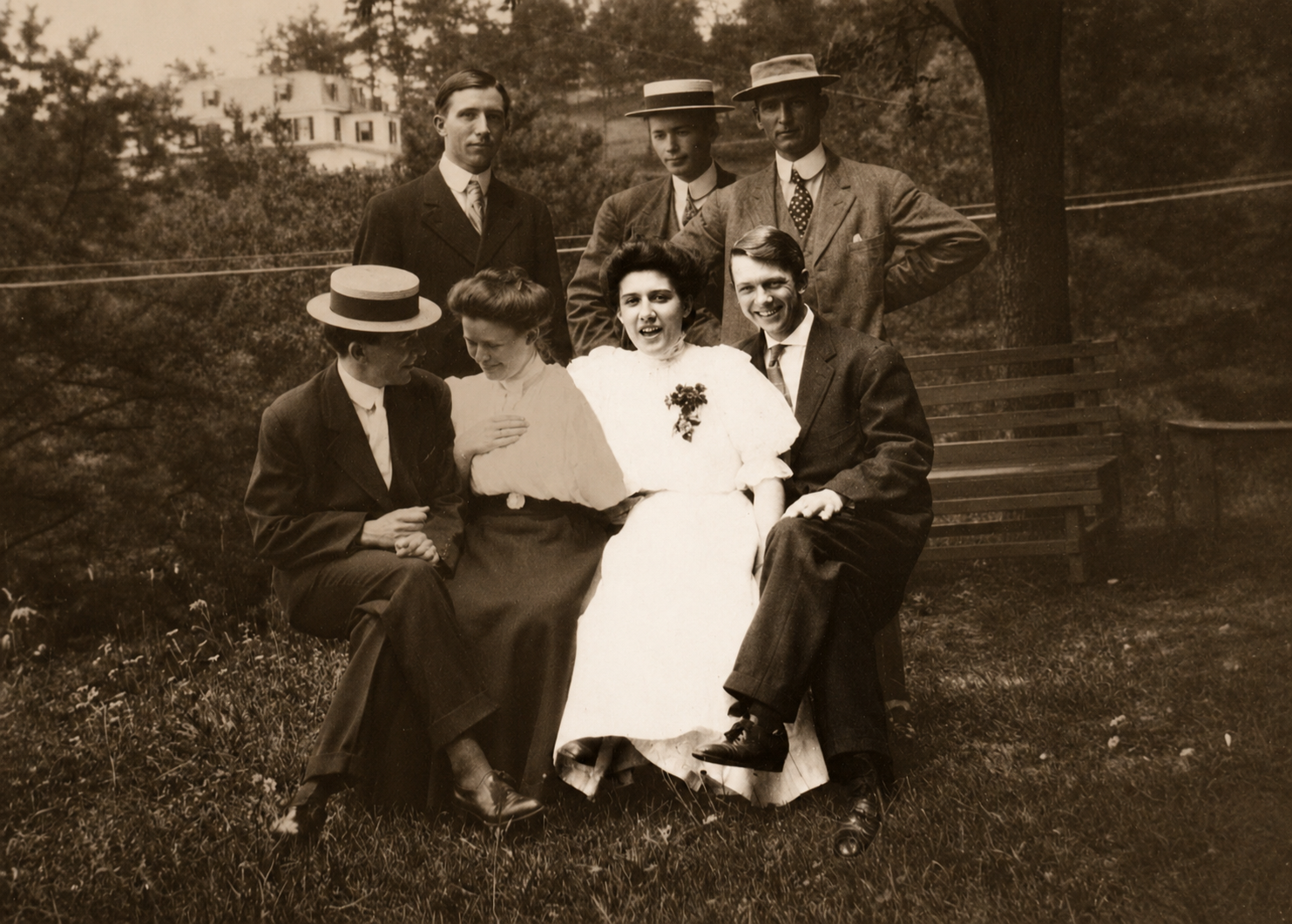 A vintage black-and-white photo of seven people outdoors. Three men are standing in the back, dressed in suits and hats. Two women and one man are seated in front, with the woman in the center smiling and wearing a white dress with a flower boutonniere, possibly indicating a wedding. The other woman is wearing a light blouse and dark skirt, and the seated man is in a suit. The background includes trees, a wooden bench, and a house on a hill.