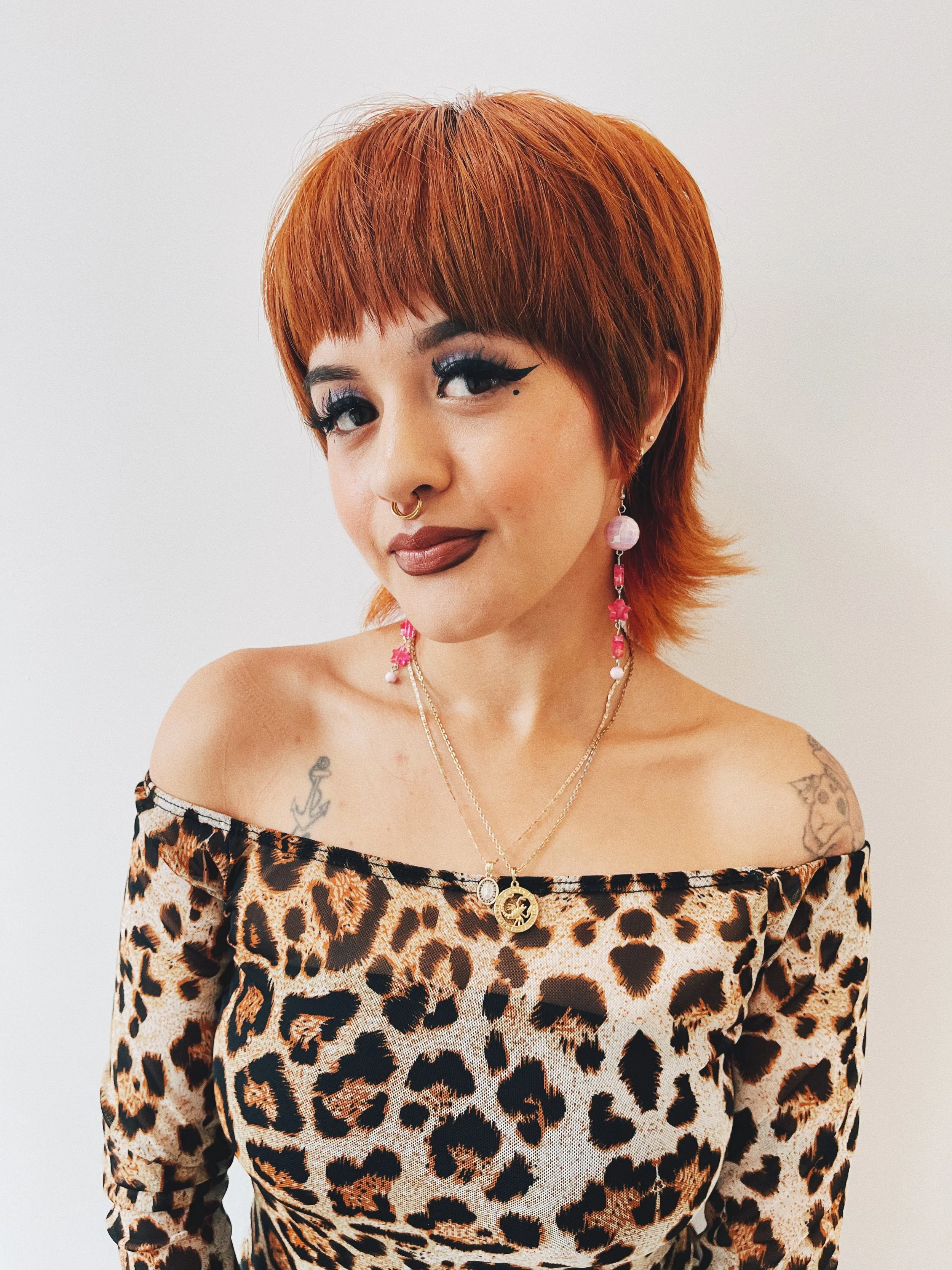 Briana Guedea, a hairstylist at The Ministry Salon in Studio City, Los Angeles, CA with red hair, wearing leopard print top, pink earrings, and necklaces, against a white background.