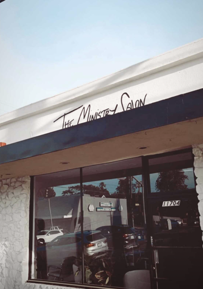 The storefront of The Ministry Salon with large glass windows and a stone wall on the side, reflecting parked cars and trees outside.