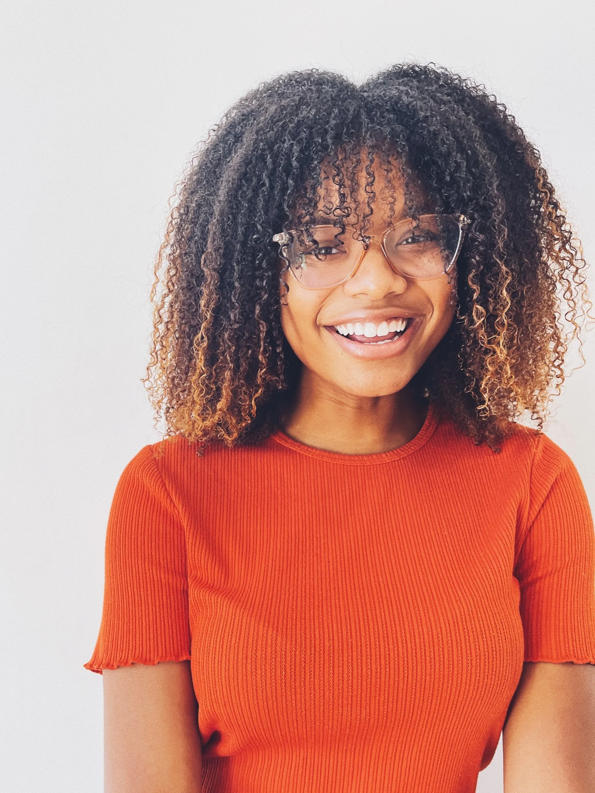 Harmoni Clawson, a hairstylist at The Ministry Salon in Studio City, Los Angeles, CA smiling, wearing glasses and an orange shirt with curly hair against a plain background.