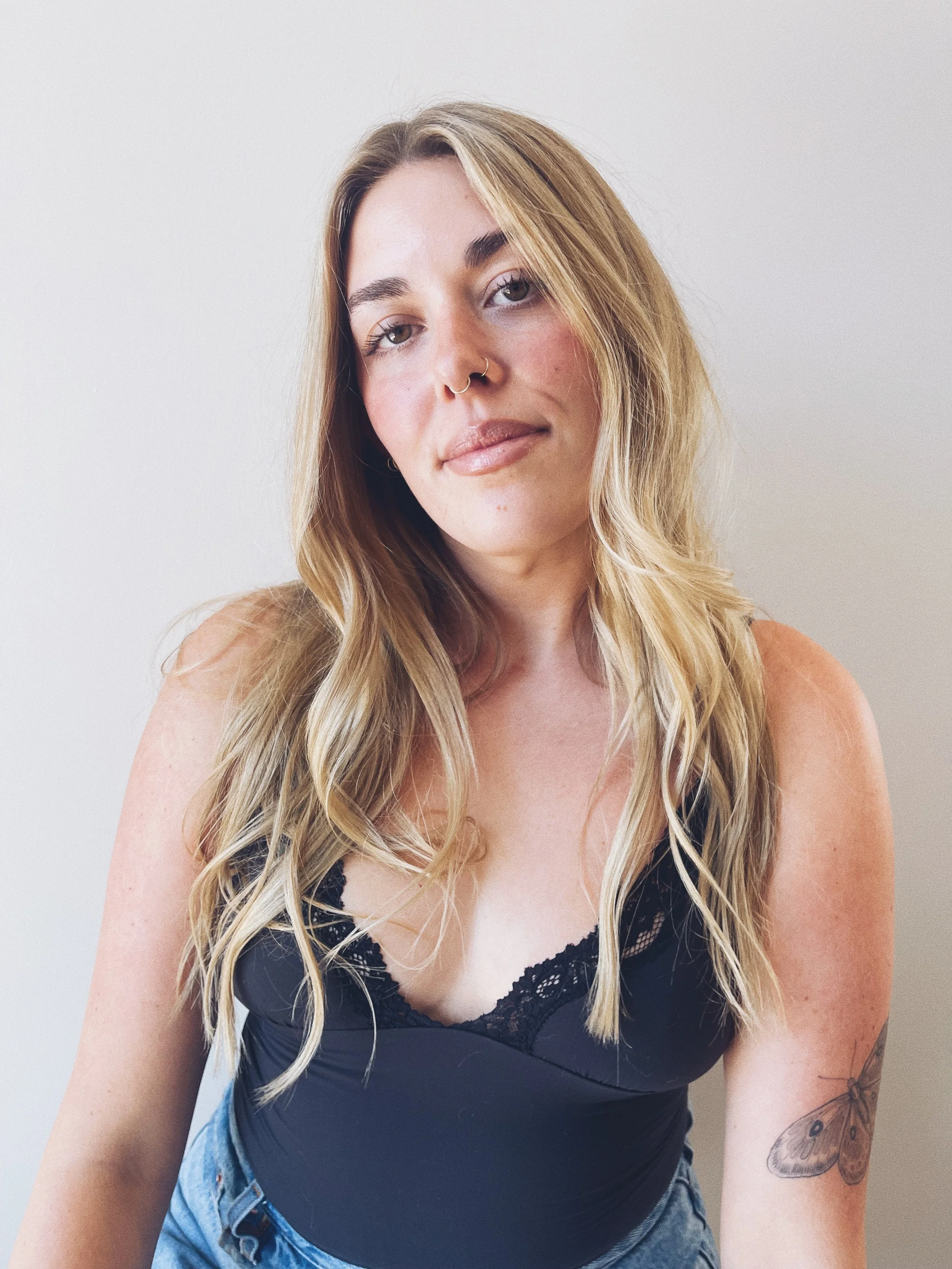 Sab Bartolone, a hairstylist at The Ministry Salon in Studio City, Los Angeles CA with long, wavy blonde hair, wearing a black lace-trimmed top, looking at the camera with a slight smile against a plain light-colored background.
