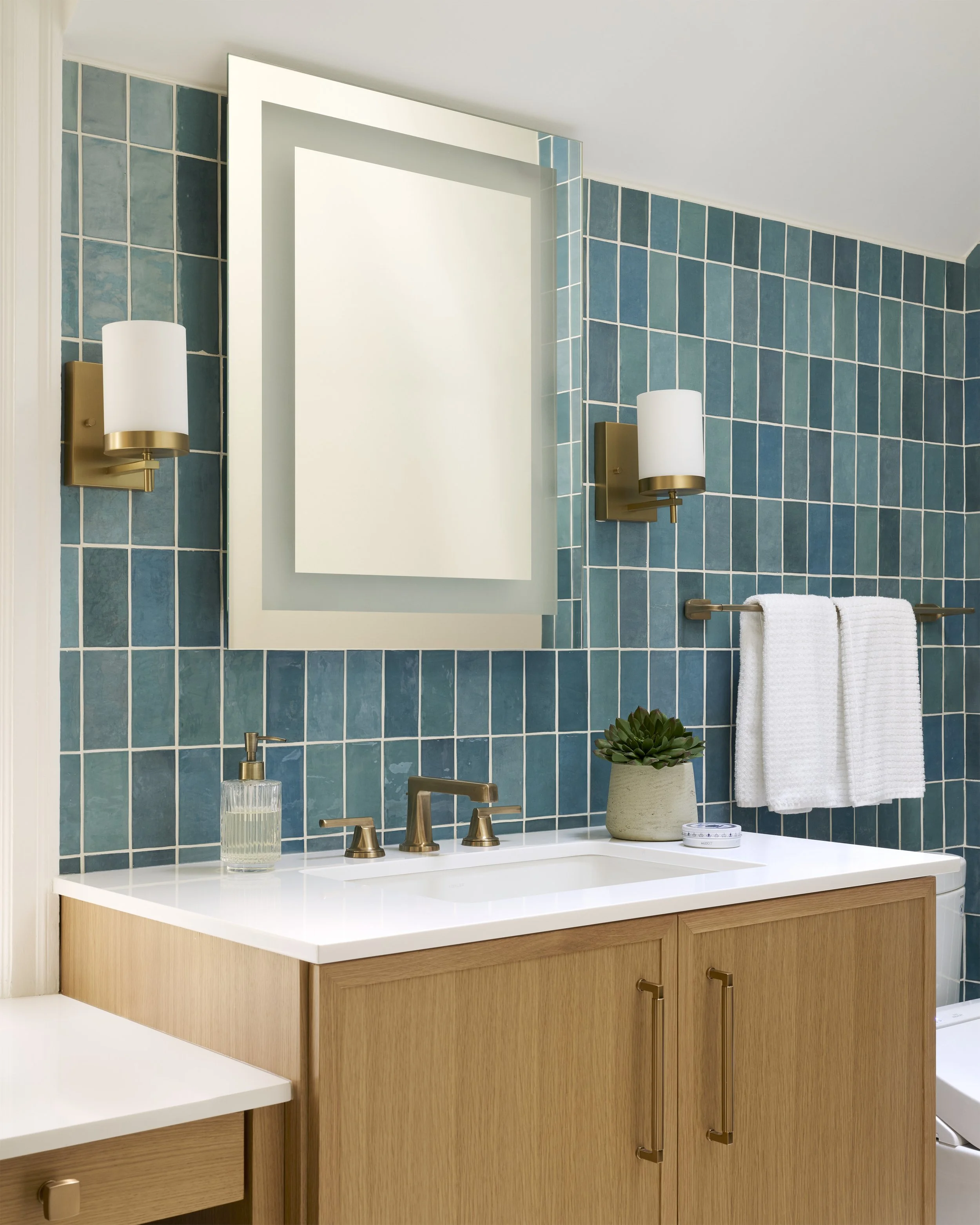transitional luxe colorful primary  bathroom with light wood vanity and white countertop, brass sconces, lighted medicine cabinet, vertical blue green tiles, slanted walls