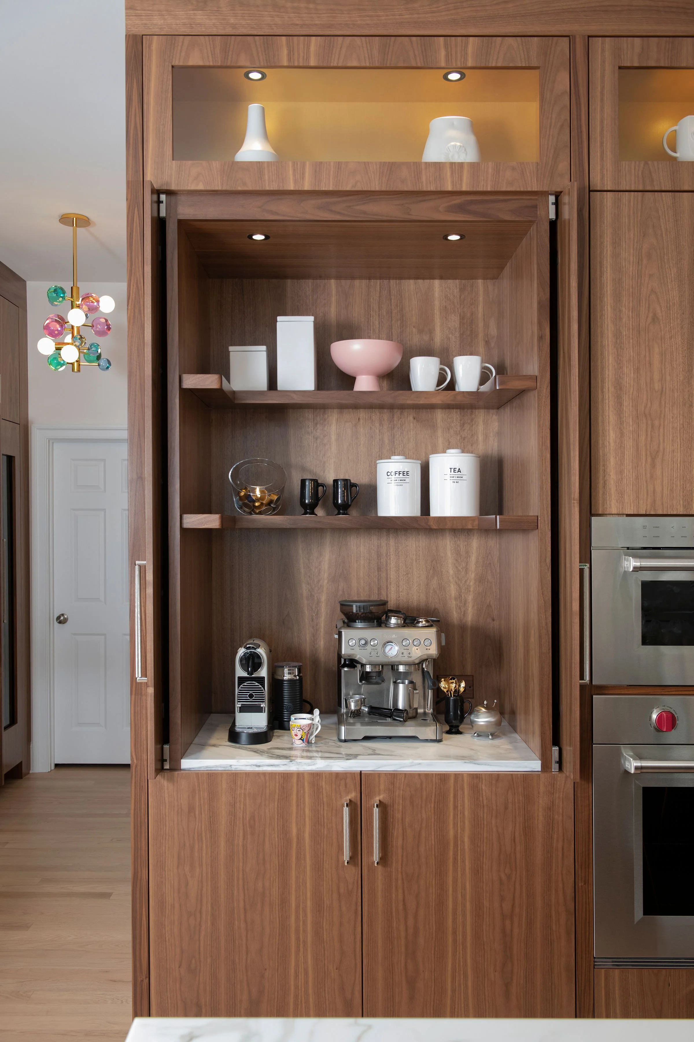 custom coffee bar in contemporary and bright kitchen with custom white and wood cabinets, white island, natural stone countertops, pendants, light boxes, gold zip drain, wolf range, double oven, coffee compartment, hidden microwave,  hardwood floors,