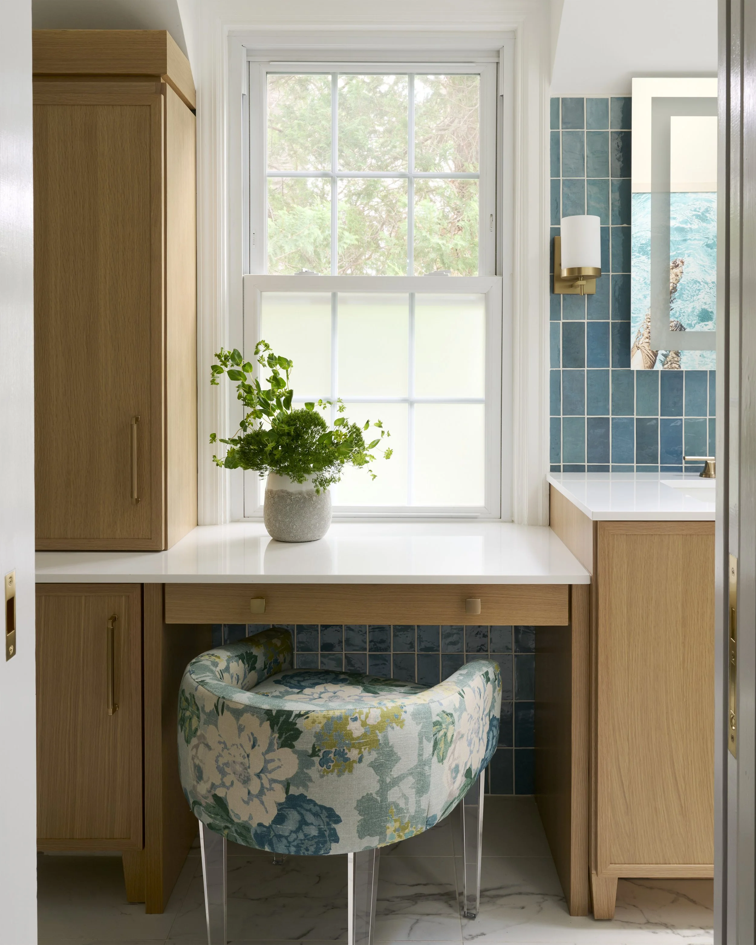 transitional luxe colorful primary  bathroom with light wood vanity and white countertop, brass sconces, lighted medicine cabinet, vertical blue green tiles, slanted walls, large scale marble shower tile, upholstered vanity chair