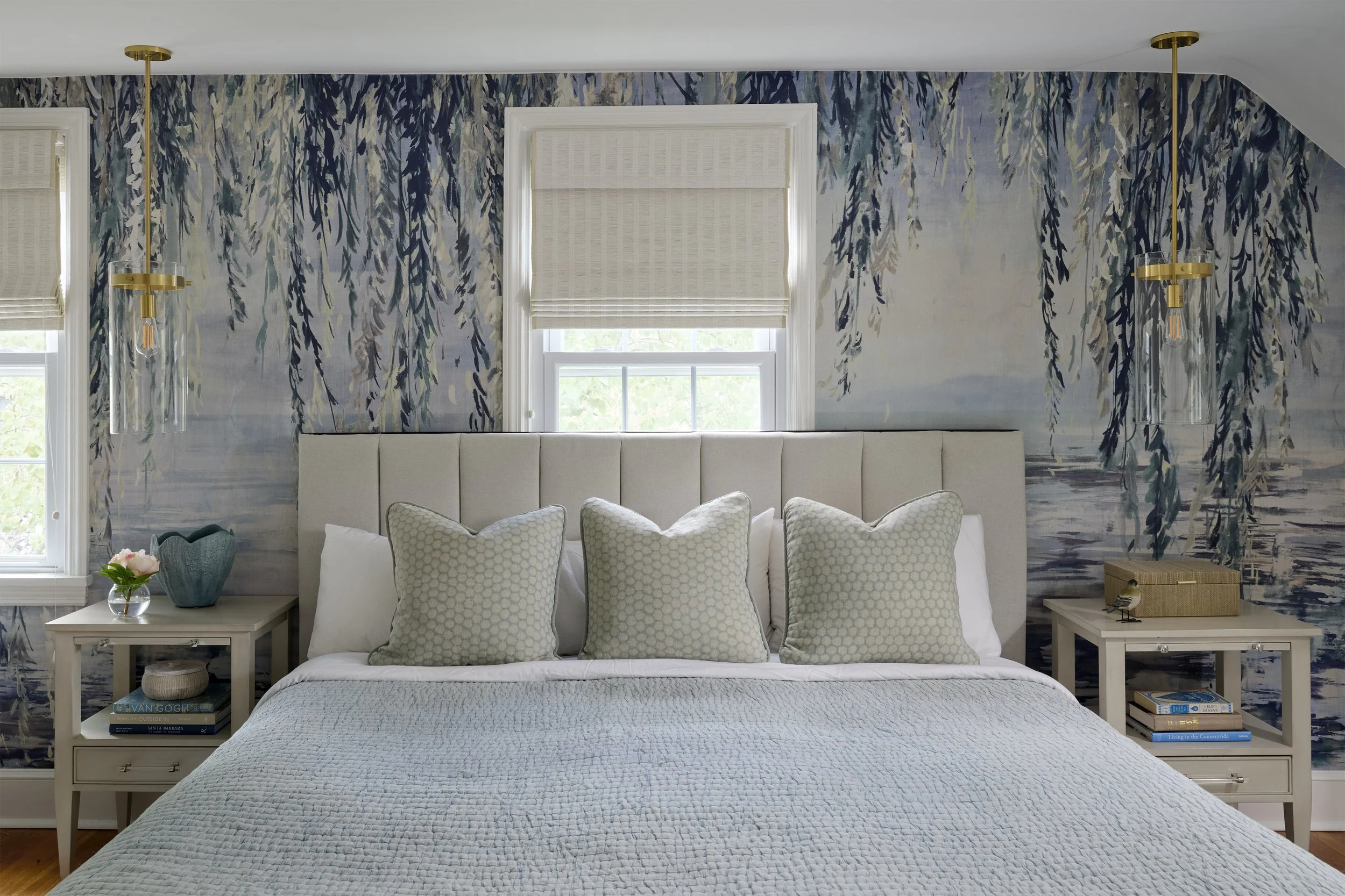 transitional luxe soothing primary bedroom with slanted ceiling, upholstered headboard, hanging clear glass pendants, three pillows, soft neutral blue bedding, and willow nature wallpaper