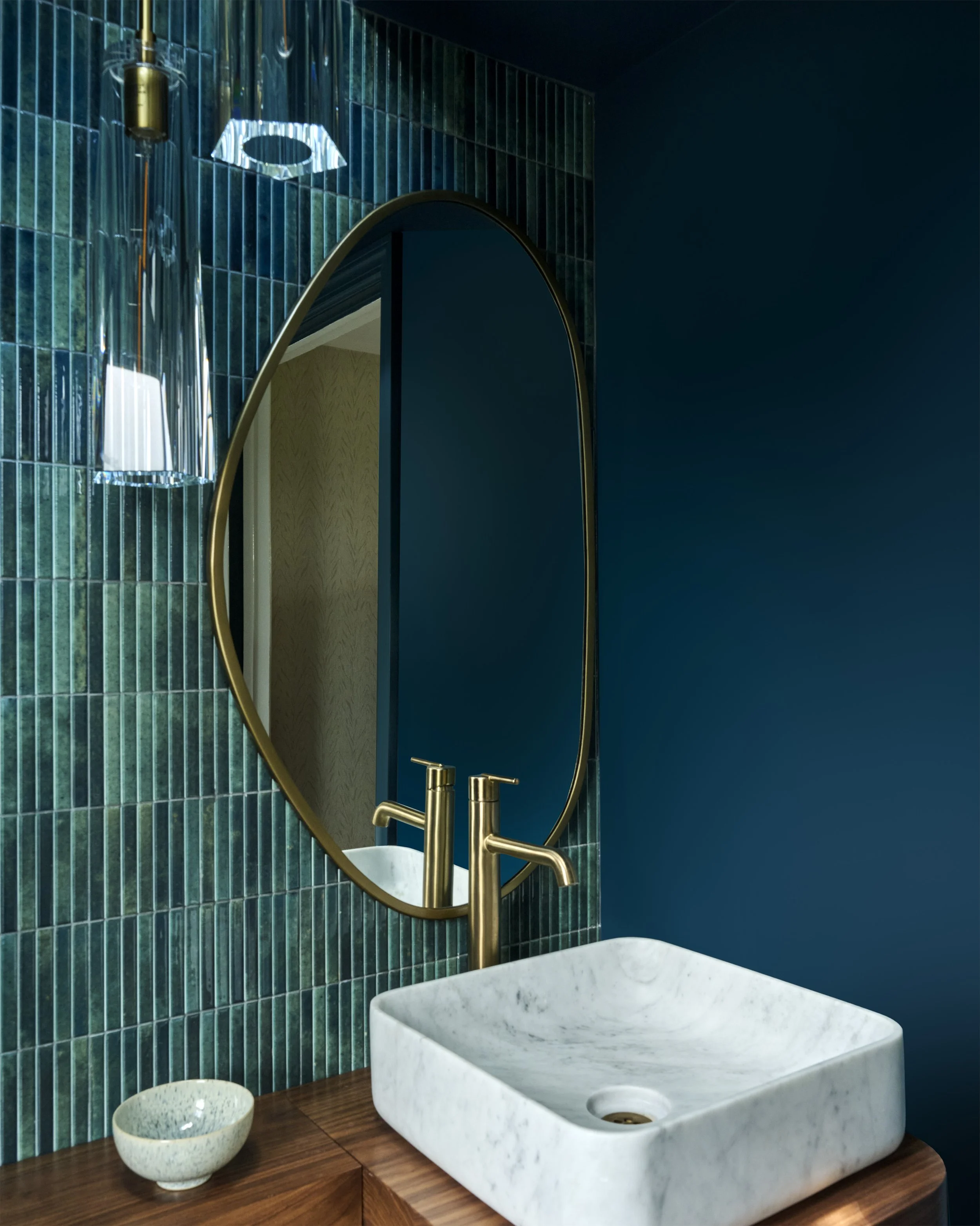 transitional rich moody luxe colorful powder room with organic brass mirror, hanging clear pendants, asymmetrical composition, blue green vertical tiles, marble vessel sink, wooden counter