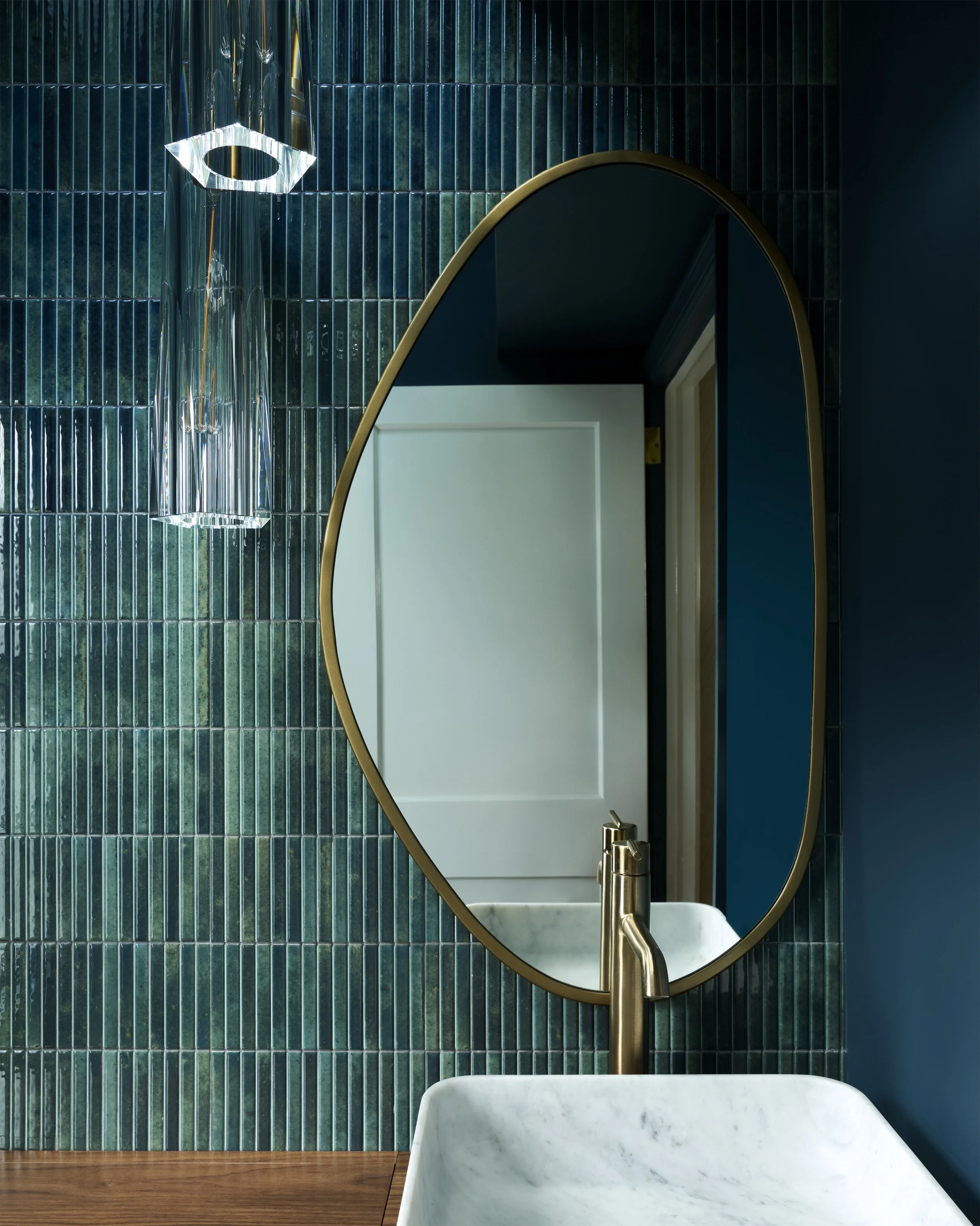 transitional rich moody luxe colorful powder room with organic brass mirror, hanging clear pendants, asymmetrical composition, blue green vertical tiles, marble vessel sink, wooden counter