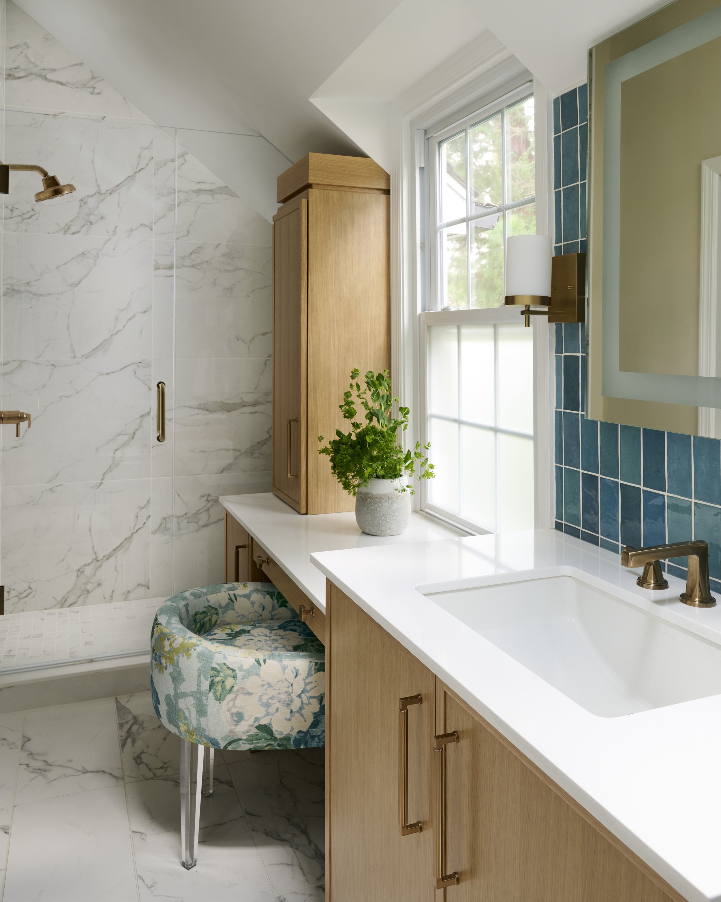 transitional luxe colorful primary  bathroom with light wood vanity and white countertop, brass sconces, lighted medicine cabinet, vertical blue green tiles, slanted walls, large scale marble shower tile, upholstered vanity chair