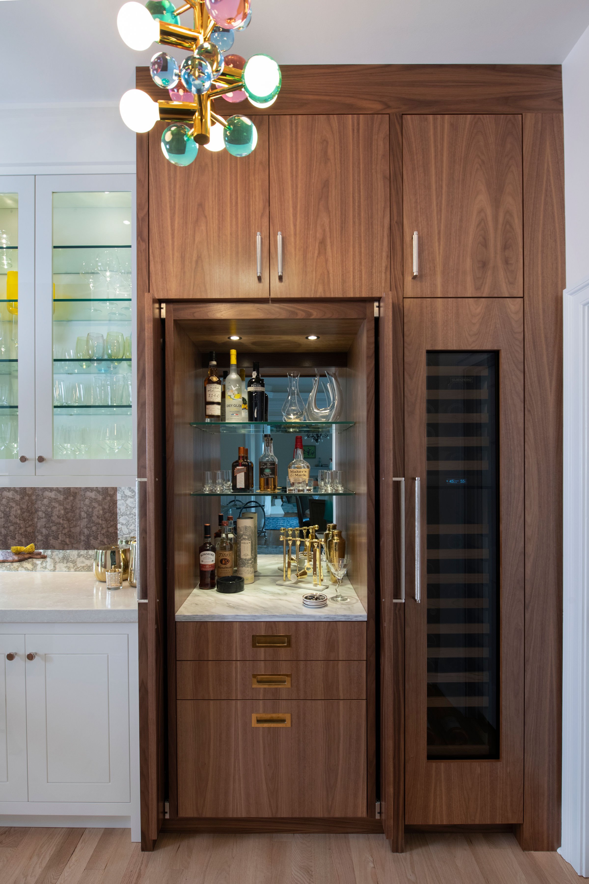 custom wine bar in contemporary and bright kitchen with custom white and wood cabinets, white island, natural stone countertops, pendants, light boxes, gold zip drain, wolf range, double oven, coffee compartment, hidden microwave,  hardwood floors,  