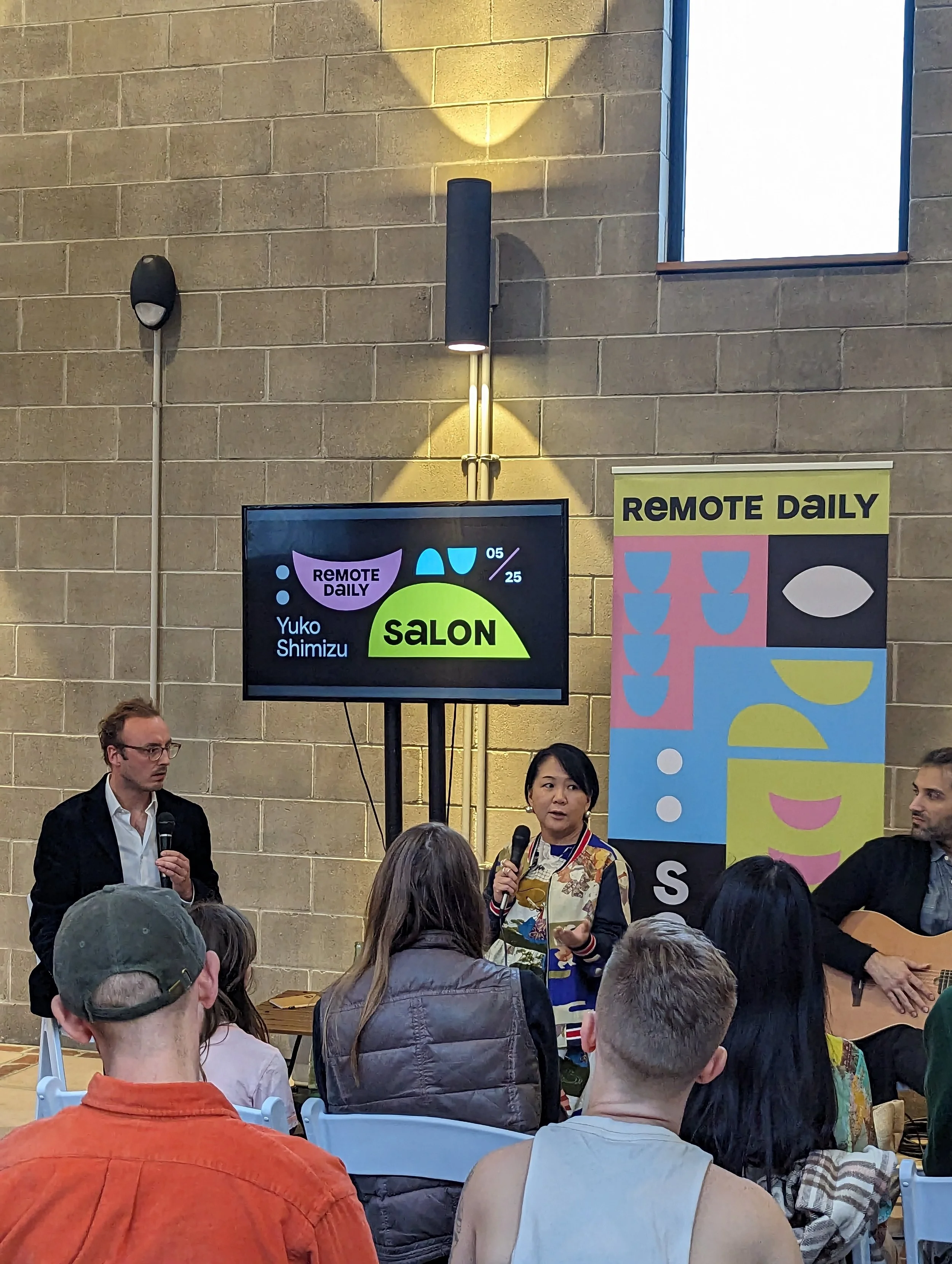 People gathered at an indoor event. A woman is speaking into a microphone, and a man is standing nearby. A large screen displays the words 'REMOTE DAILY' and some graphics. There are banners and a group of audience members watching.