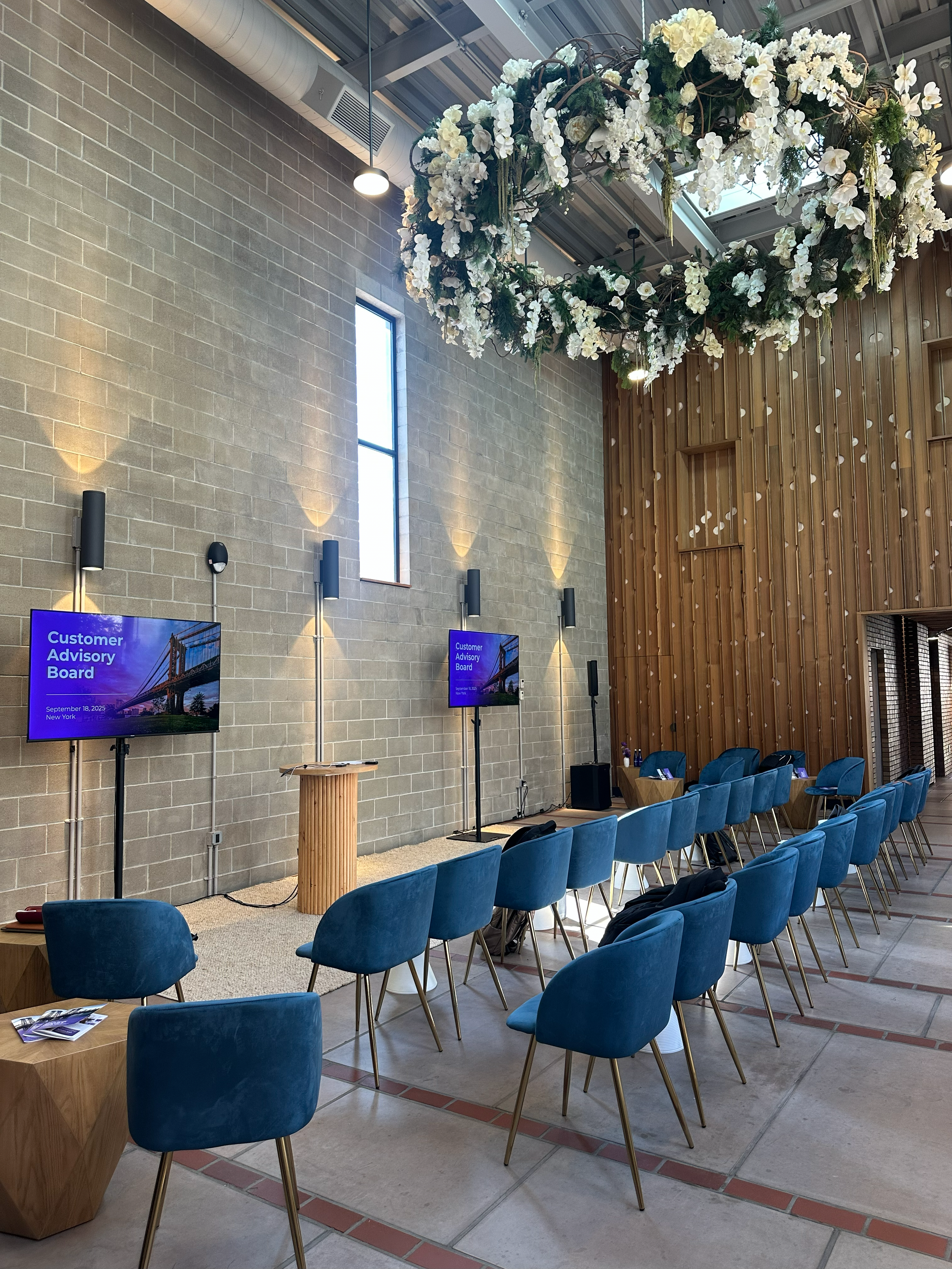 A conference room setup with rows of blue cushioned chairs facing a stage area with a wooden speaker's podium and two large screens displaying a presentation titled 'Customer Advisory Board' scheduled for September 18, 2025, in New York. The room features a high ceiling with modern lighting, a large window, brick and wooden wall accents, and decorative floral arrangements hanging from the ceiling.