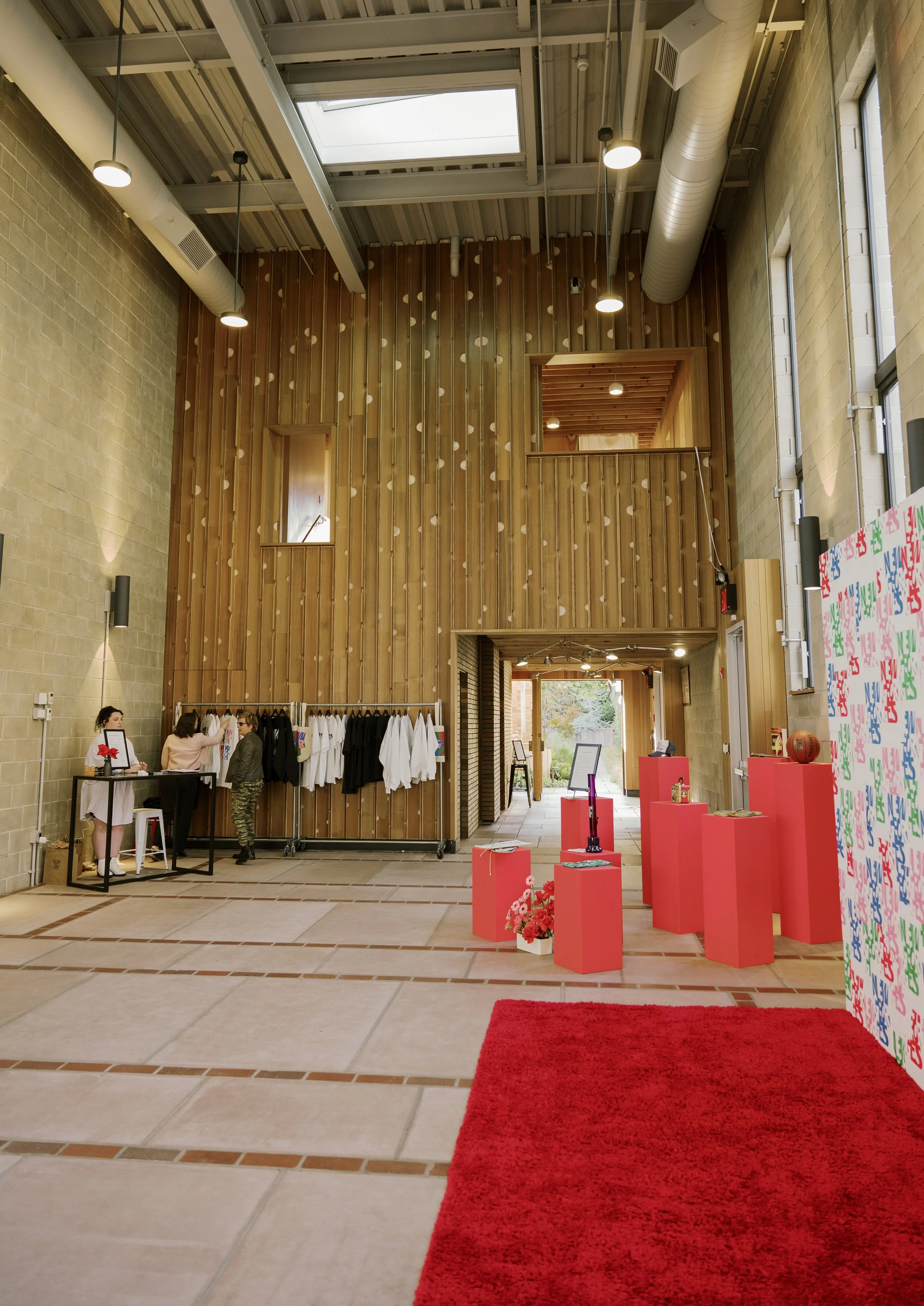 Interior of a modern building with high ceiling, large windows, and a red carpet on the floor. There are display tables with flowers and collectibles, such as a basketball, and decorative stands. A clothing rack with white and black shirts is on the left, and people are browsing and chatting near the display area.