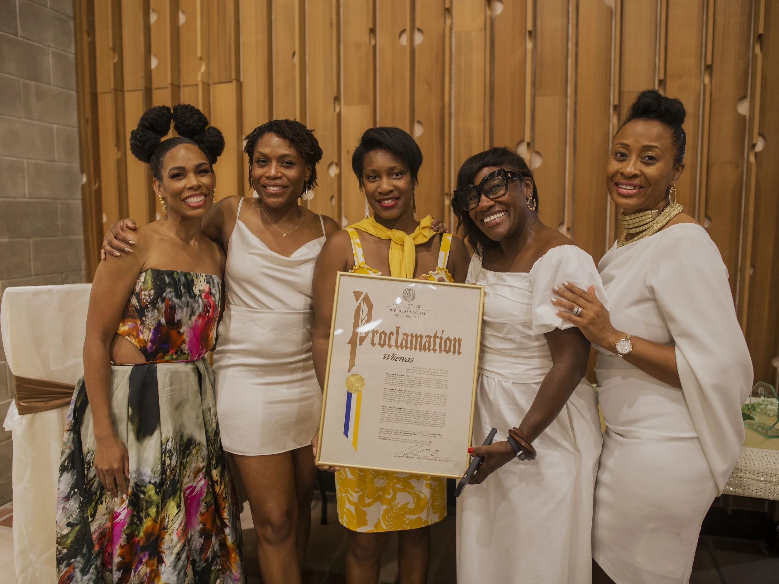 Five women standing together at an event, smiling, with one holding a framed certificate or award, in front of a wooden wall in the indoor space at Carroll Hall in Brooklyn, NY.