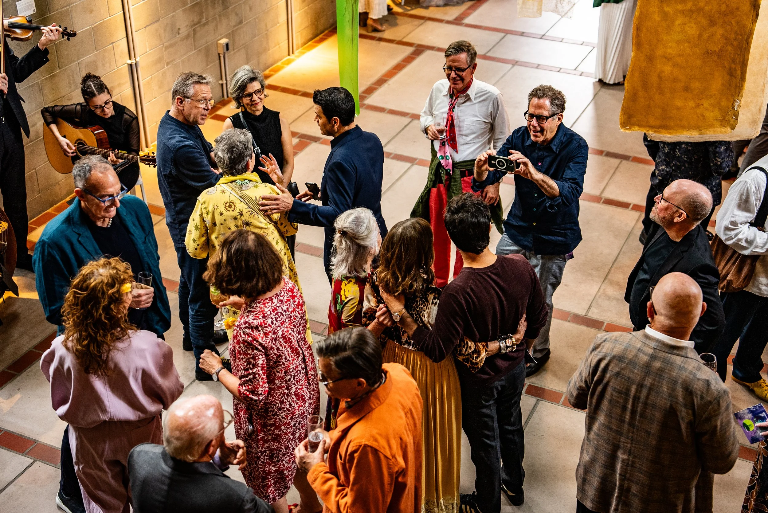 People gathered in a room, some dancing, some playing musical instruments, and others taking photos, at a social event or celebration.