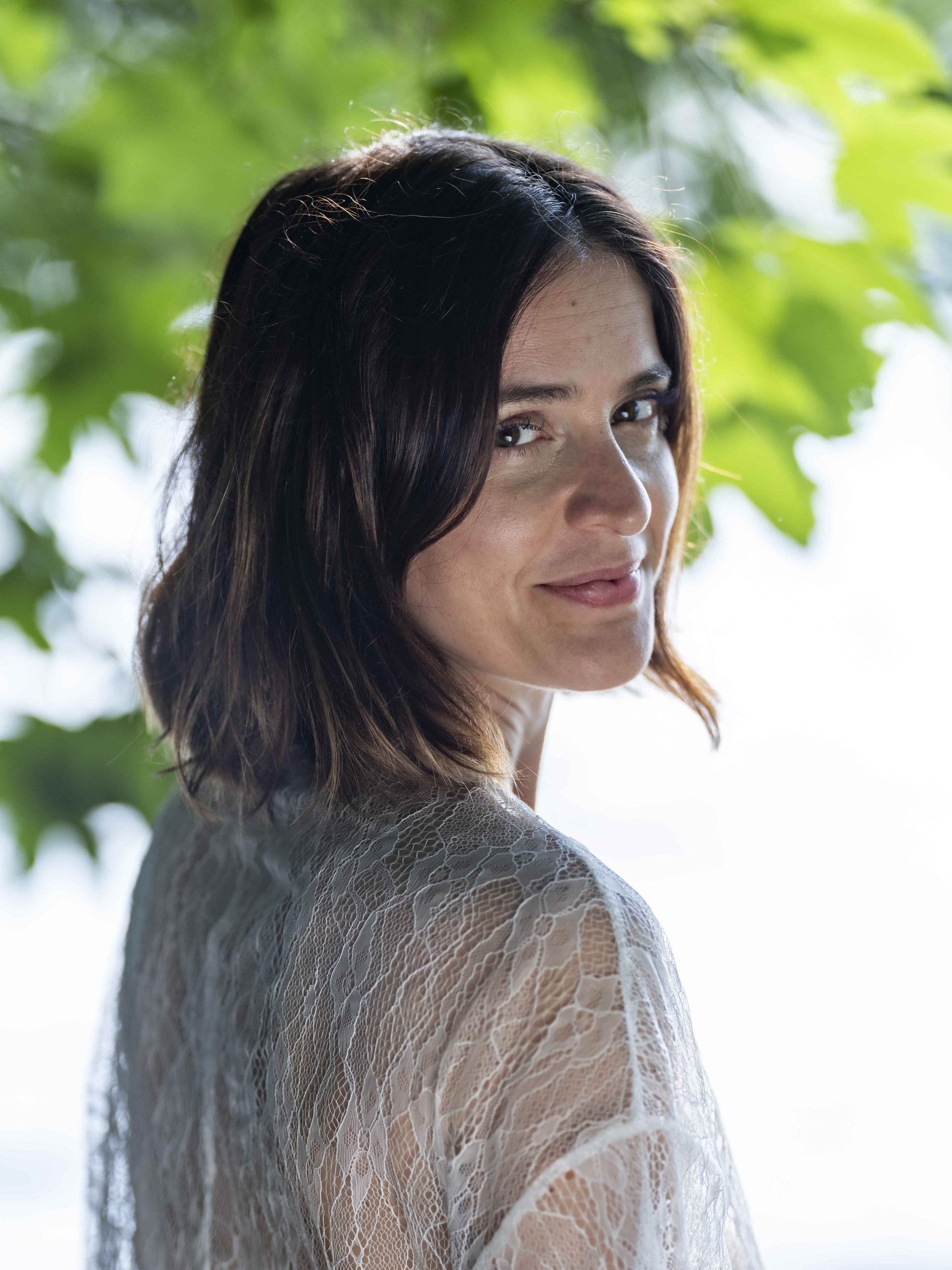 A woman with dark, shoulder-length wavy hair looks over her shoulder, wearing a lace top with a blurred green leafy background.