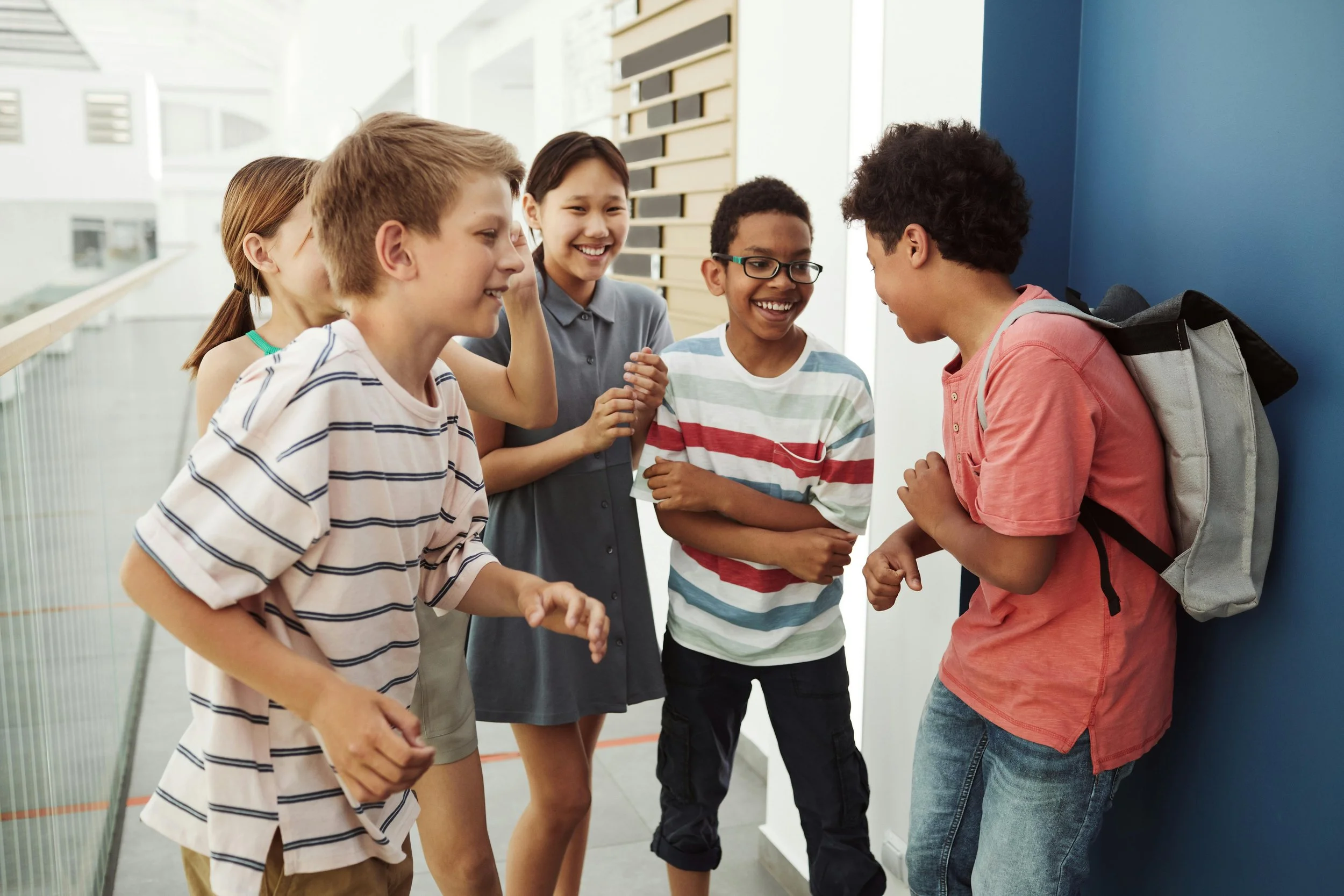 Group of five middle school children laughing and talking in a school hallway, one boy leaning against a wall, wearing a red shirt and backpack, four girls standing nearby.