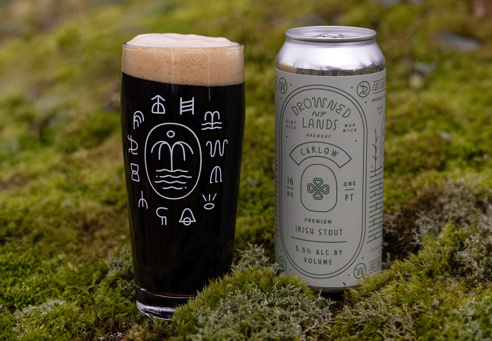 A glass filled with dark stout beer with a light foam head, next to a tall aluminum can of Irish stout beer with a pale green label featuring various green designs and text, set on green mossy ground.