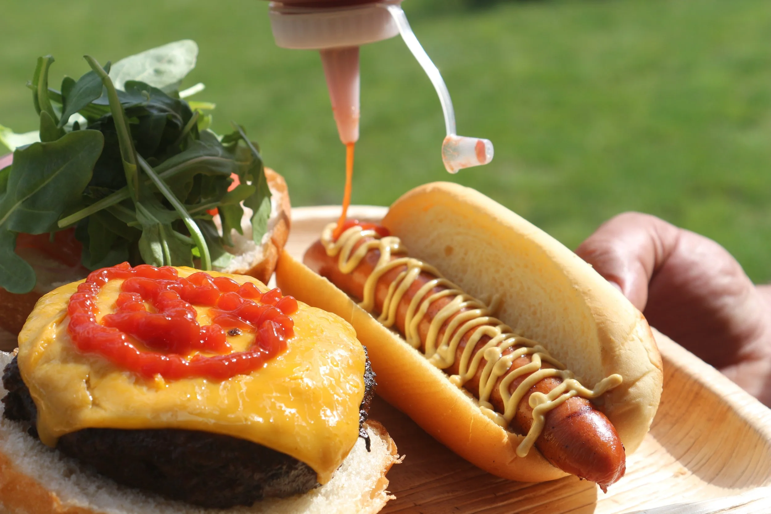 A hot dog with mustard, a cheeseburger with ketchup, and a small salad on a wooden tray, with mustard being squeezed onto the hot dog.