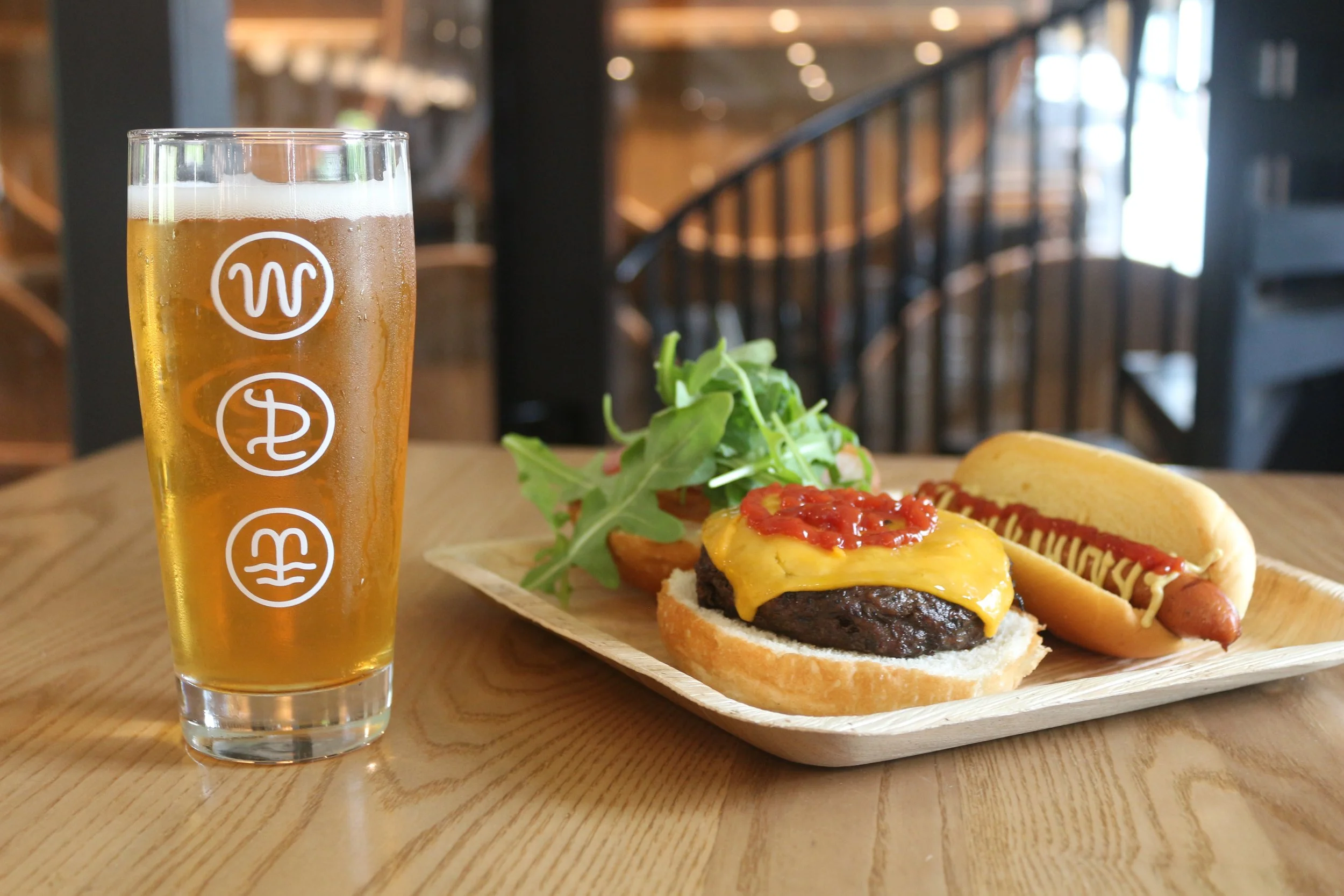 A tray with a cheeseburger with cheddar cheese, ketchup, and a hamburger patty, a hot dog with mustard and ketchup, and mixed greens on a wooden table, with a glass of beer on the side, in a restaurant with wooden interior.