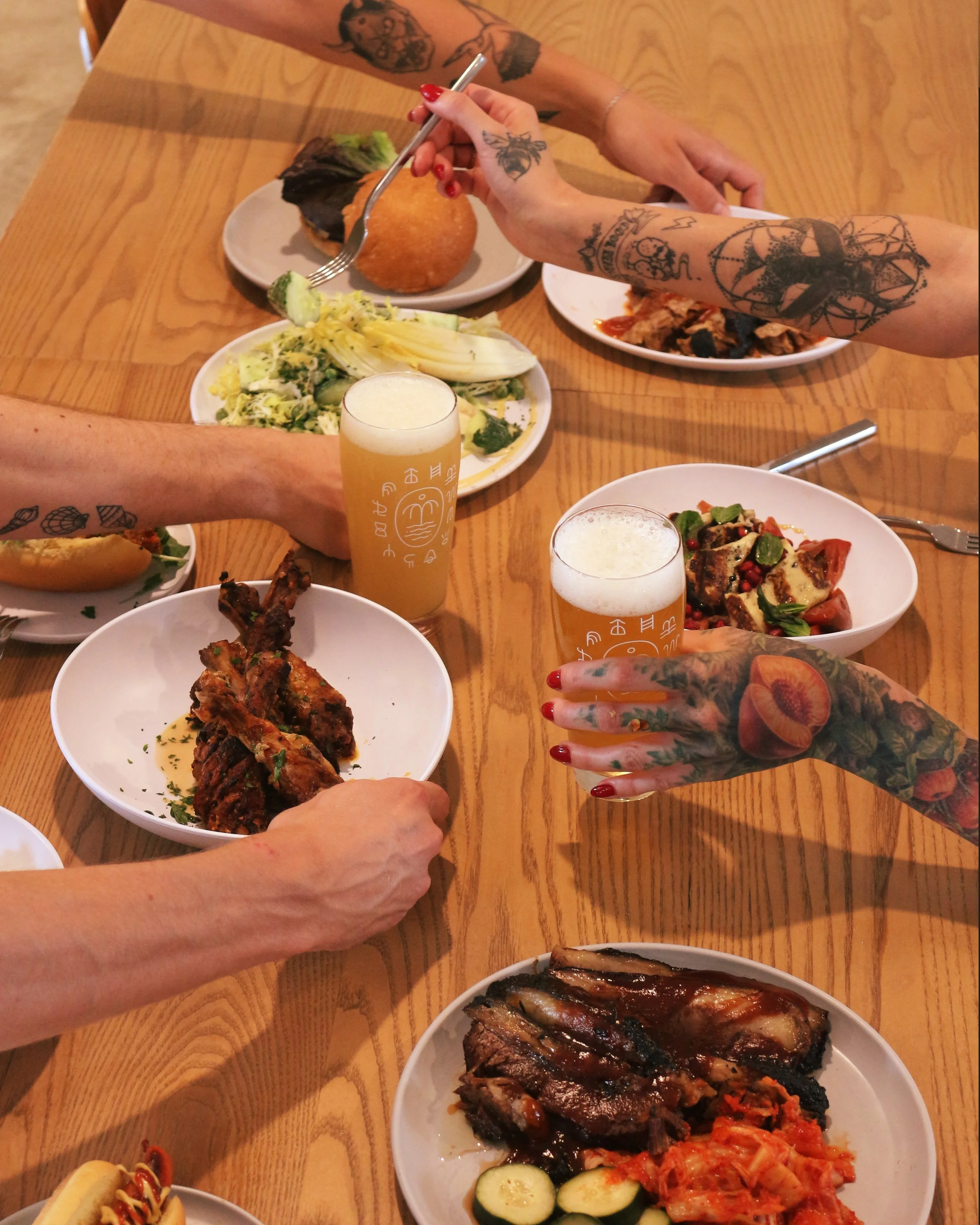 A group of people sharing a meal at a wooden table, with various dishes including salad, ribs, fried chicken, grilled vegetables, and beverages.
