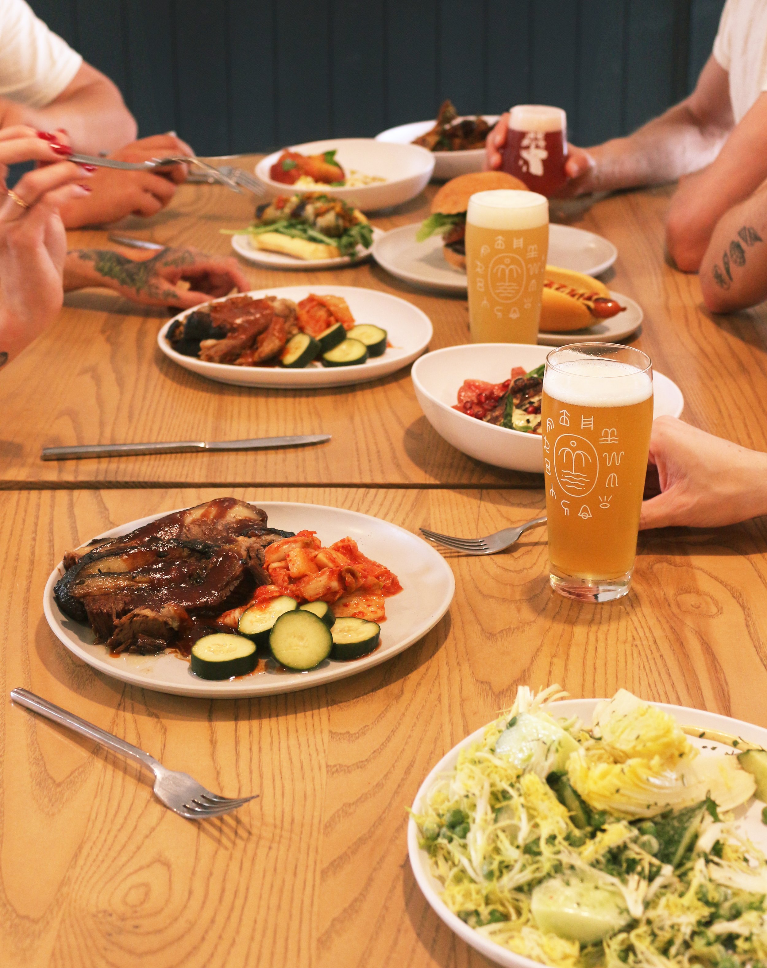 A table with various plates of food, including salads, grilled meat, vegetables, and sandwiches, with glasses of beer among people dining.
