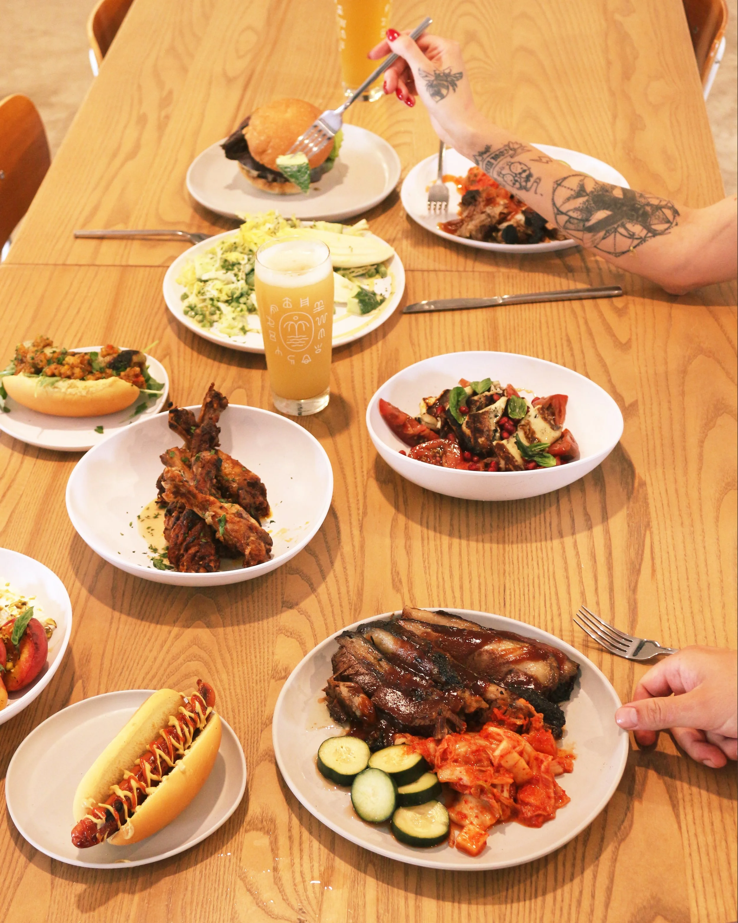 A variety of dishes on a wooden table, including ribs, kimchi, grilled vegetables, a hot dog, salad, fried chicken, burger, and drinks, with people serving and eating.