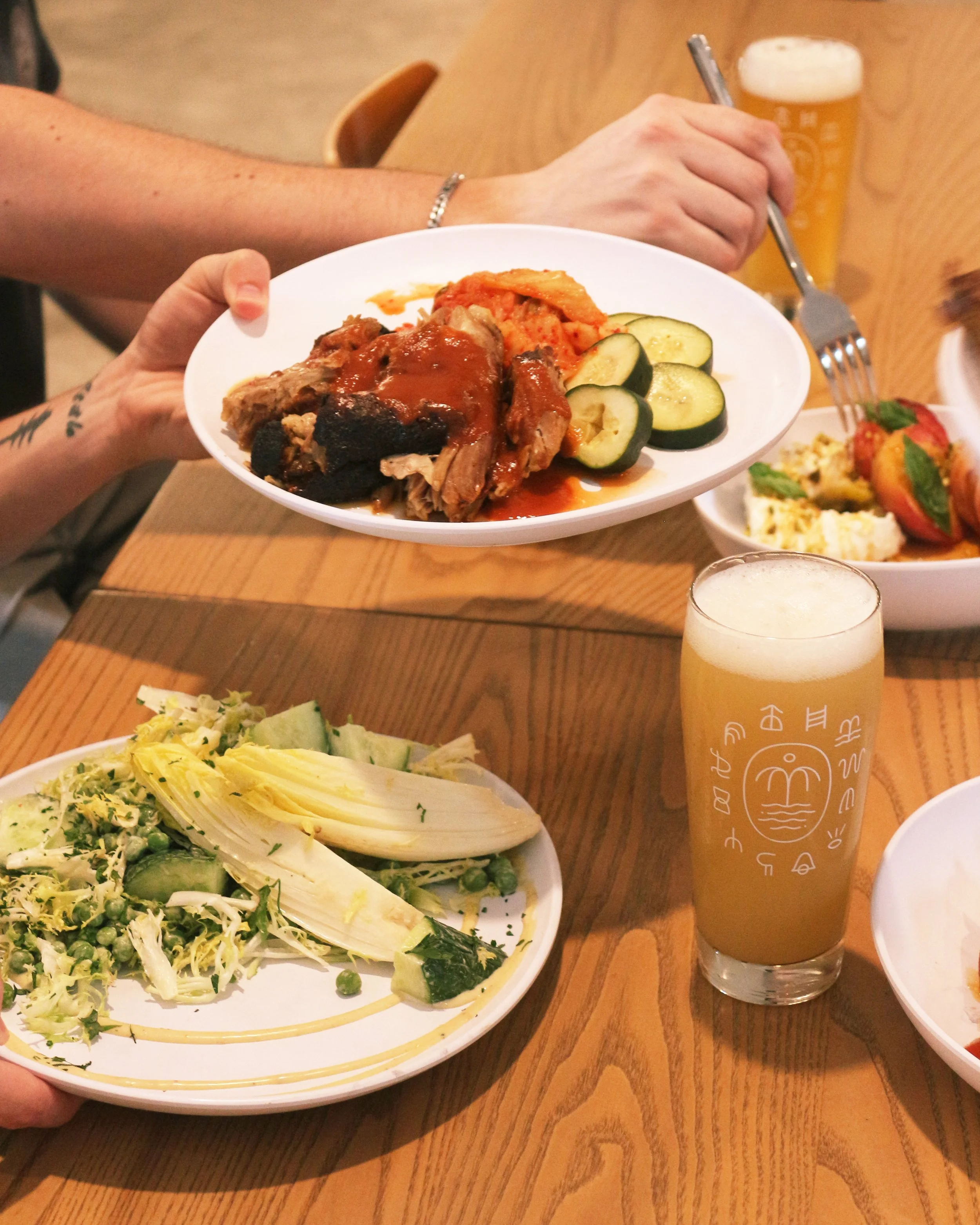 Dining scene with plates of salad, chicken with sauce, and sliced cucumbers, along with glasses of beer on a wooden table.