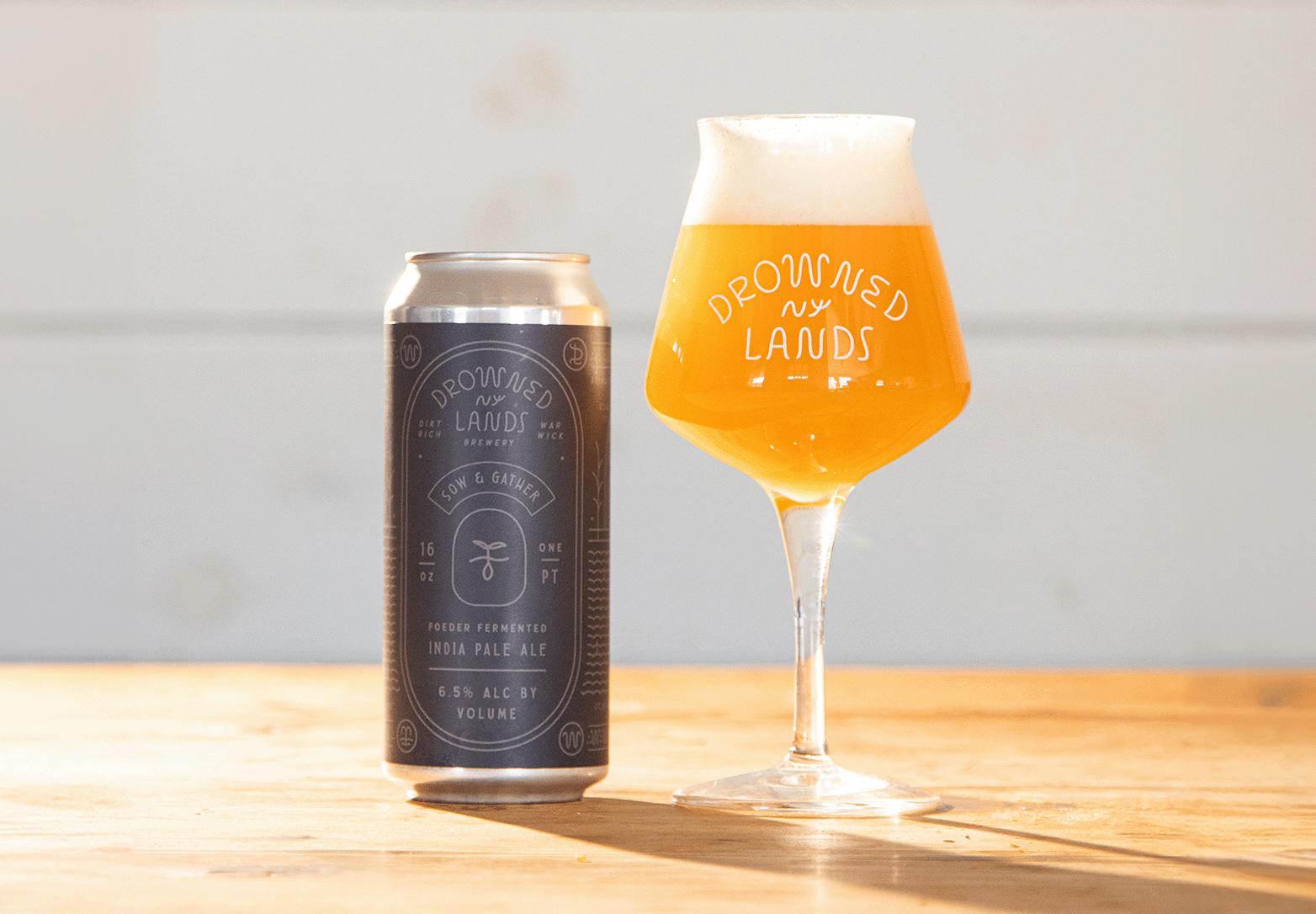 A can of Drownsed Land brewery India Pale Ale next to a tulip-shaped beer glass filled with a hazy, golden beer, both placed on a wooden surface.