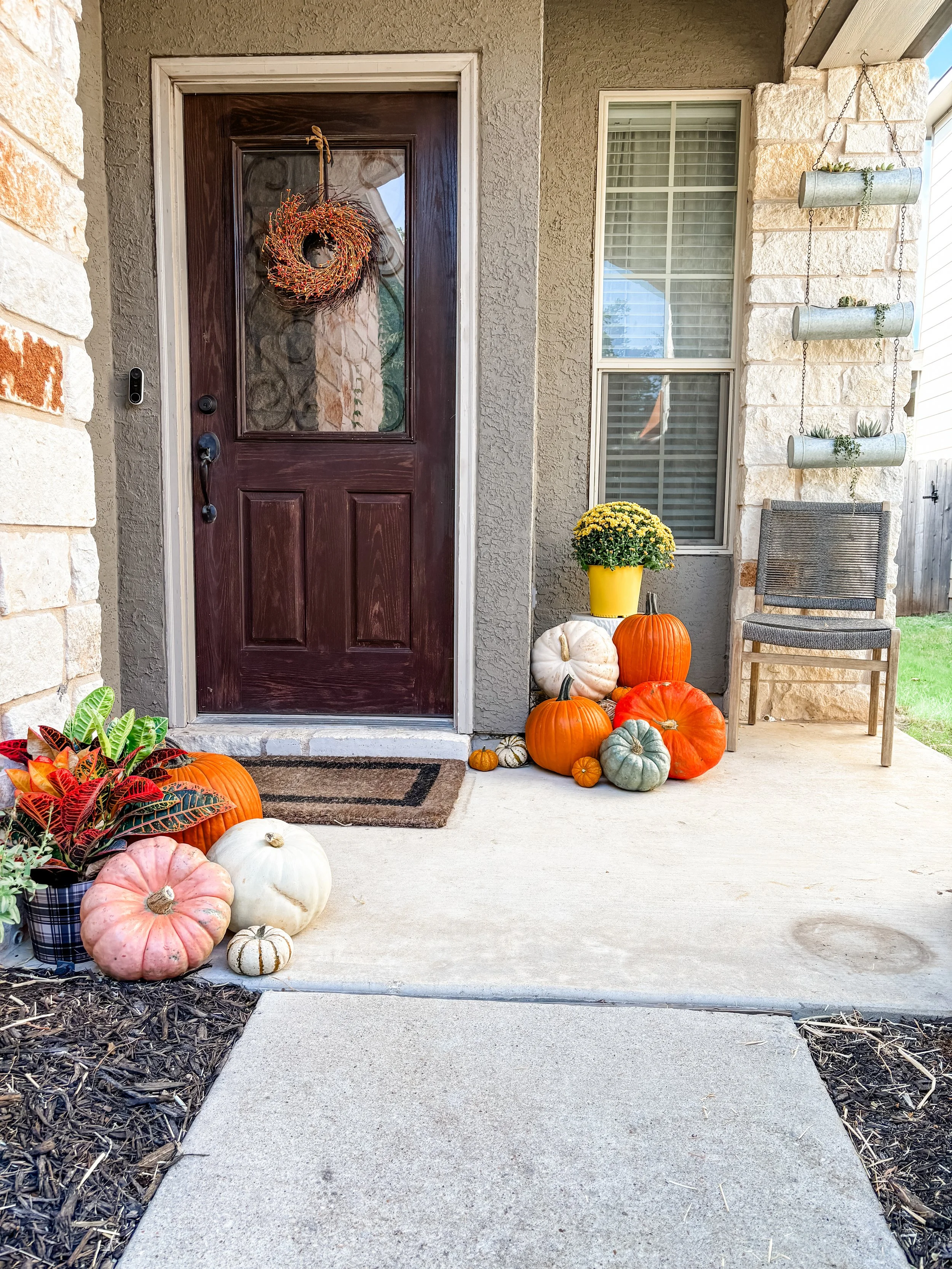 Front porch decorated for fall with pumpkins, gourds, and plants, featuring a wooden door with a wreath, a chair, and a window.