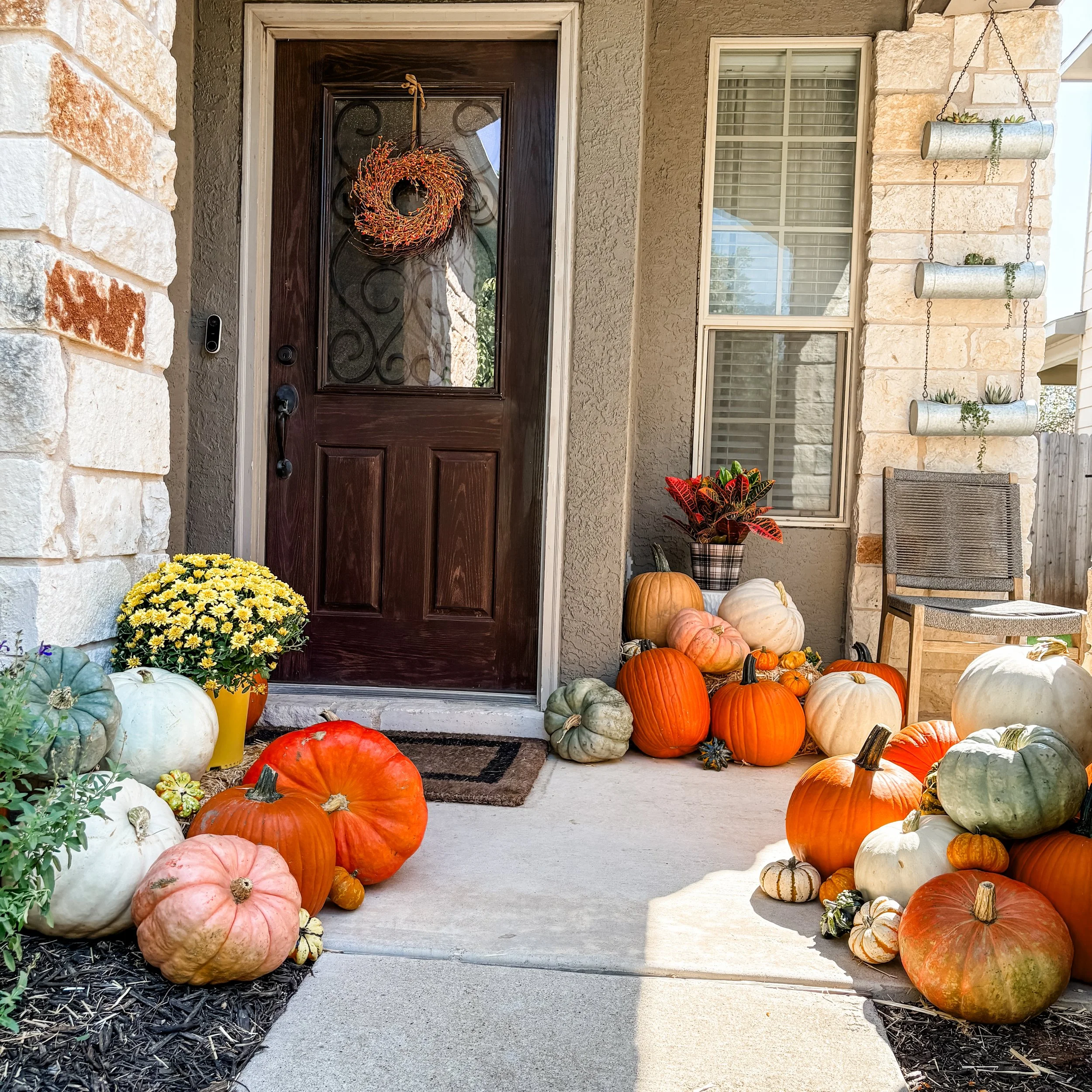 Fall harvest decorations on a porch include pumpkins of various sizes and colors, a potted plant with red and green leaves, a yellow potted chrysanthemum, and a chair with hanging planters on the wall. A wreath hangs on the door.