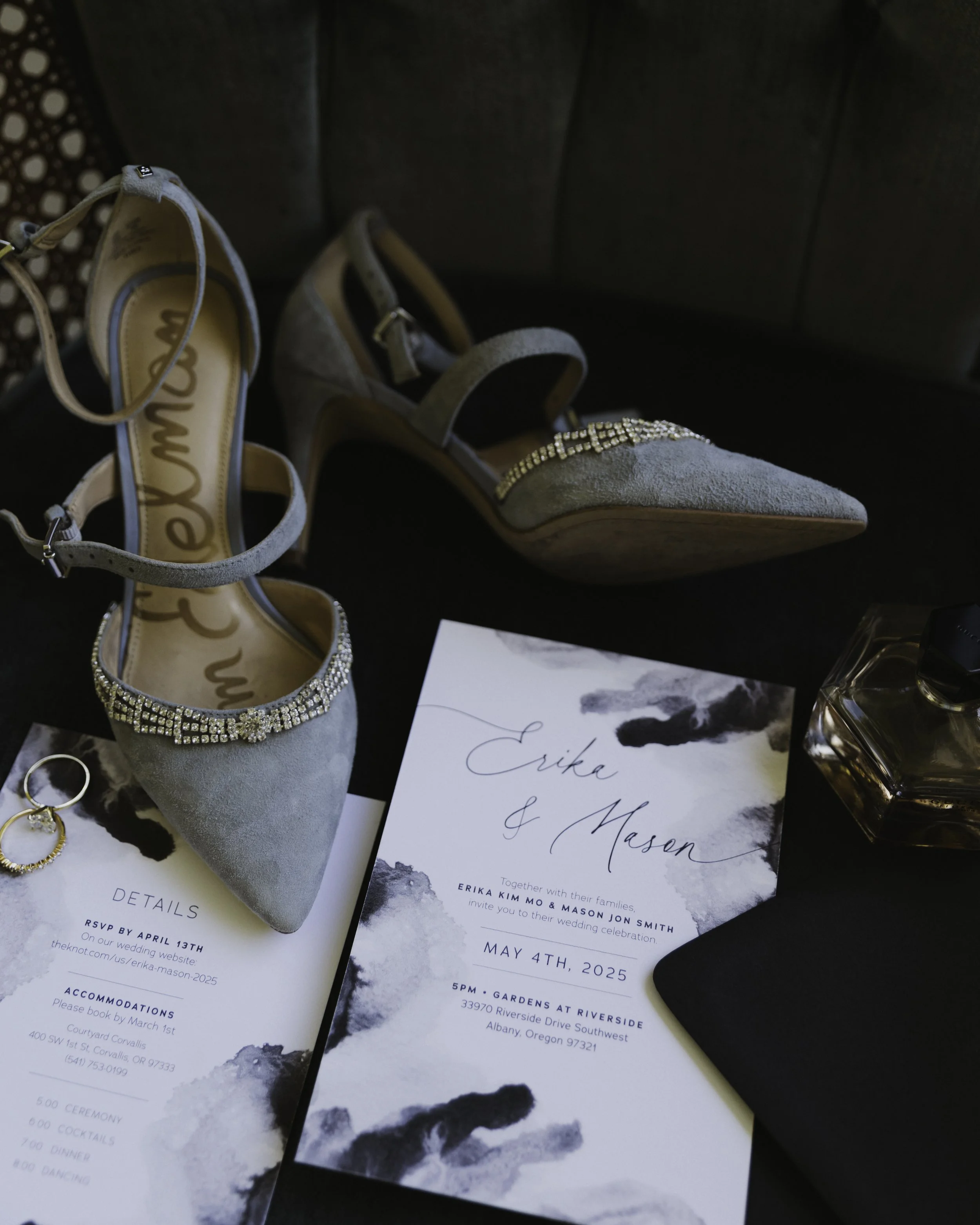 A black and white marbled watercolor wedding invitation for Erika and Mason, a second card featuring the wedding details, a perfume bottle, and a pair of gold earrings laid out on a dark surface, and a pair of gray high-heeled shoes with rhinestones.