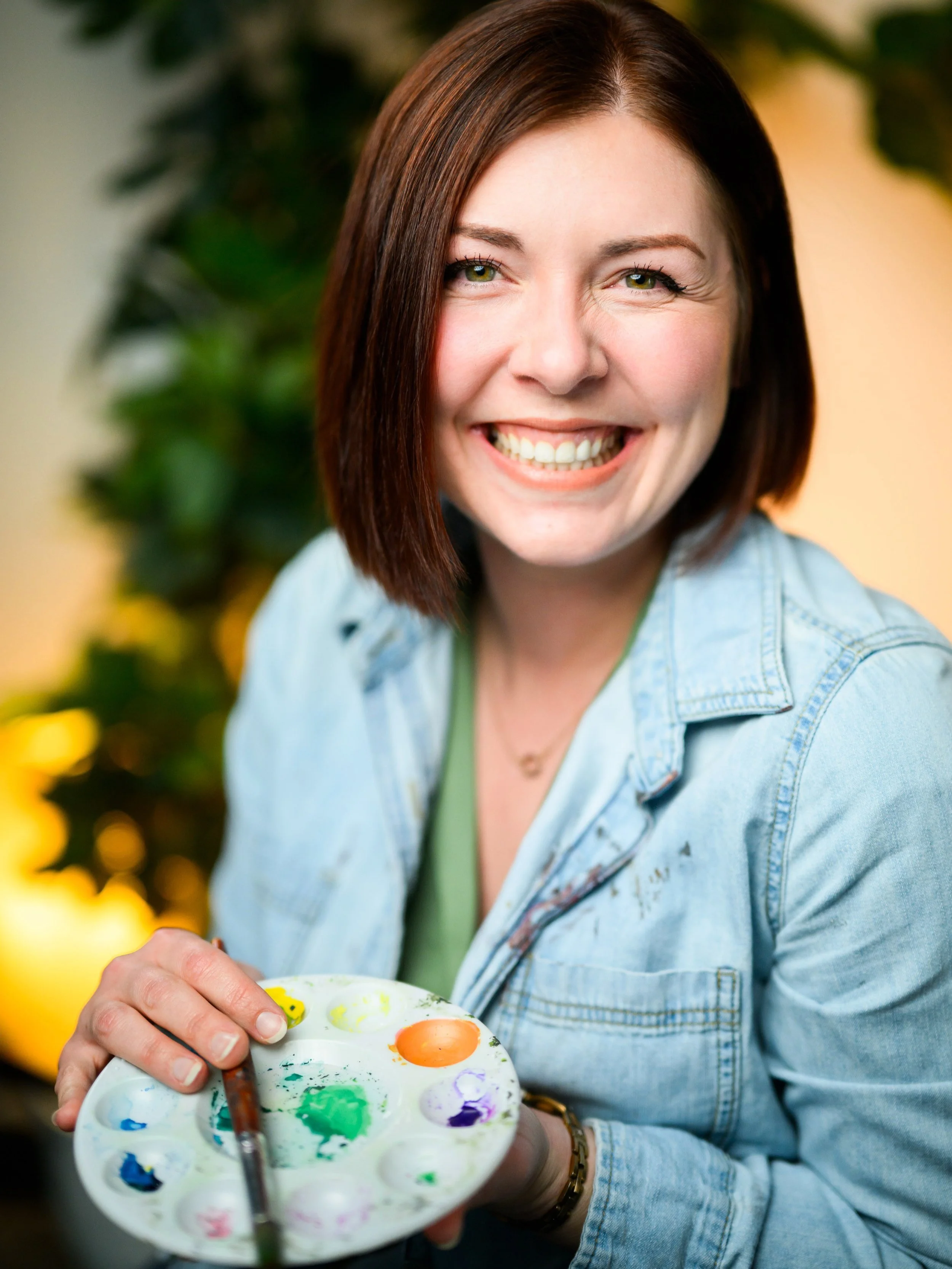 Stationery designer Michelle with shoulder-length brown hair and a bright smile is holding a paint palette with various colors and a paintbrush, with blurred green foliage in the background.