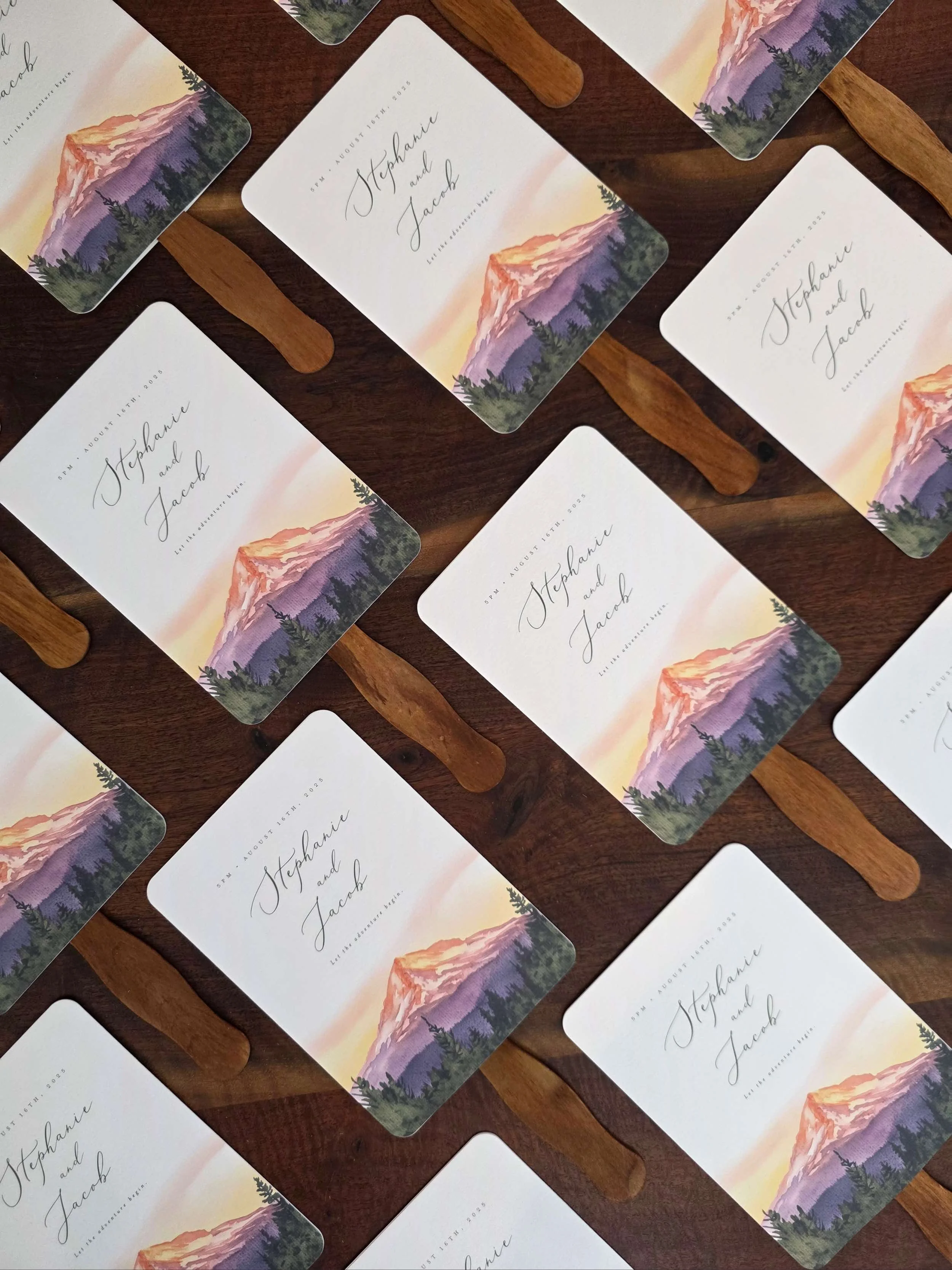 Wedding program fans with custom watercolor art of Mt Hood at sunset with pine trees with cursive typography on paper with a wooden handle