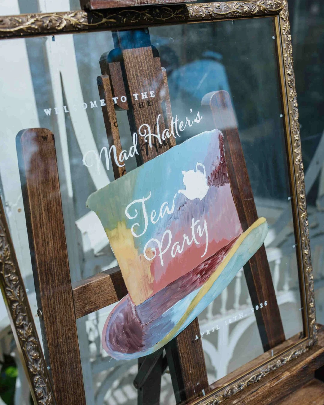 Hand-painted alternative wedding welcome sign with mad hatter's tea party text on a colorful hat painted on glass with a large antique frame.