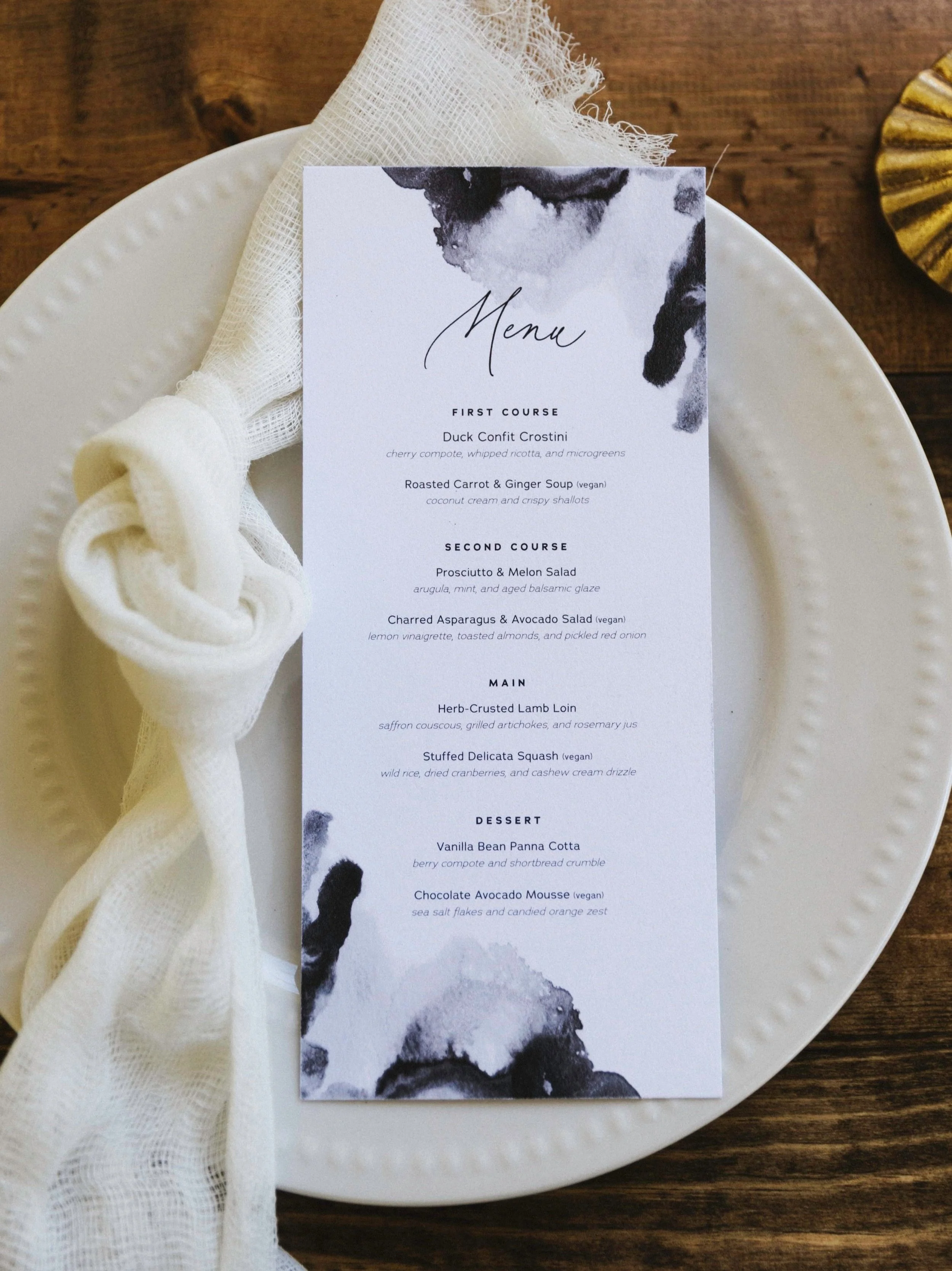 Elegant 4"x9" dinner menu with black and white watercolor art on white paper, placed on a white plate with a white cloth napkin on a wooden table.