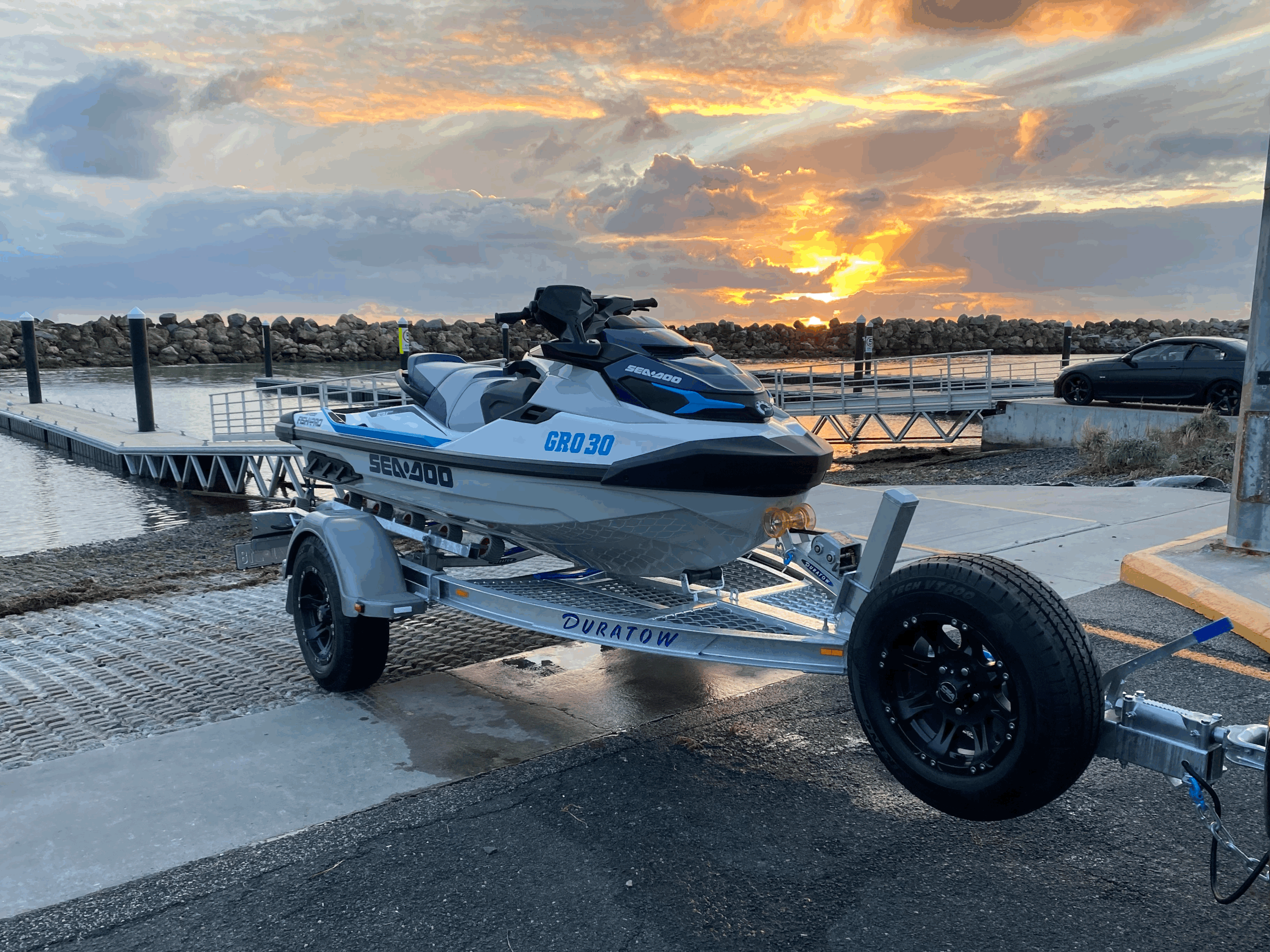 A jet ski on a trailer at a marina during sunset with clouds and a partly visible car in the background.
