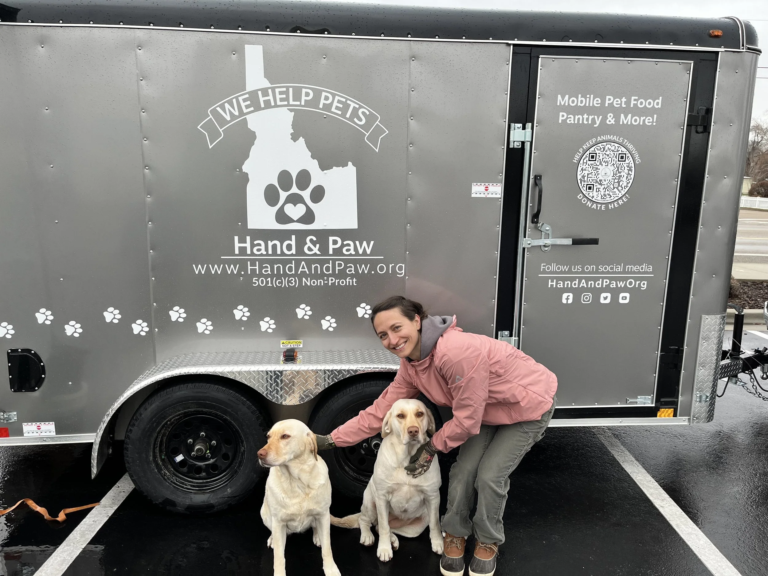 Hand and Paw mobile pet food pantry trailer with Kristina, Founder and President, standing with two beautiful yellow labs who just received some pet food and treats.