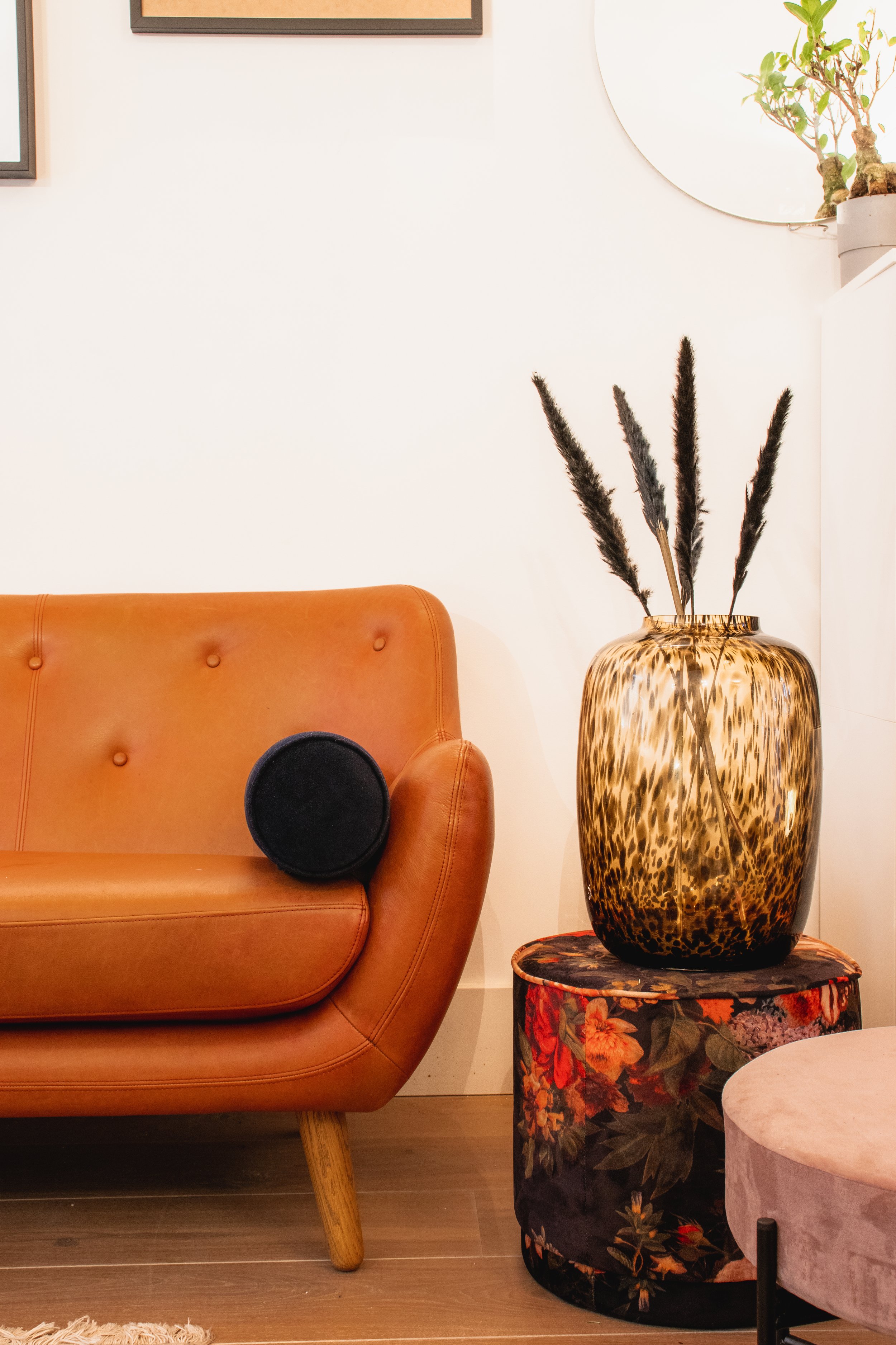 Mid-century modern living room with a brown leather sofa, black cushion, large gold-speckled vase with dark decorative branches, floral-patterned ottoman, round mirror, and a small potted plant.