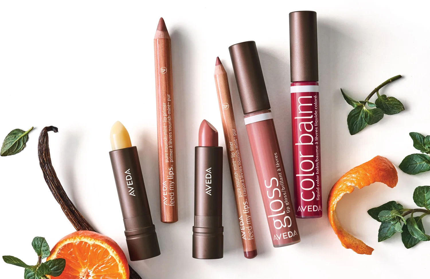 Aveda Feed My Lips lip primer, liner, lipstick, gloss and color balm laid out with a vanilla bean, a halved tangerine and fresh mint
