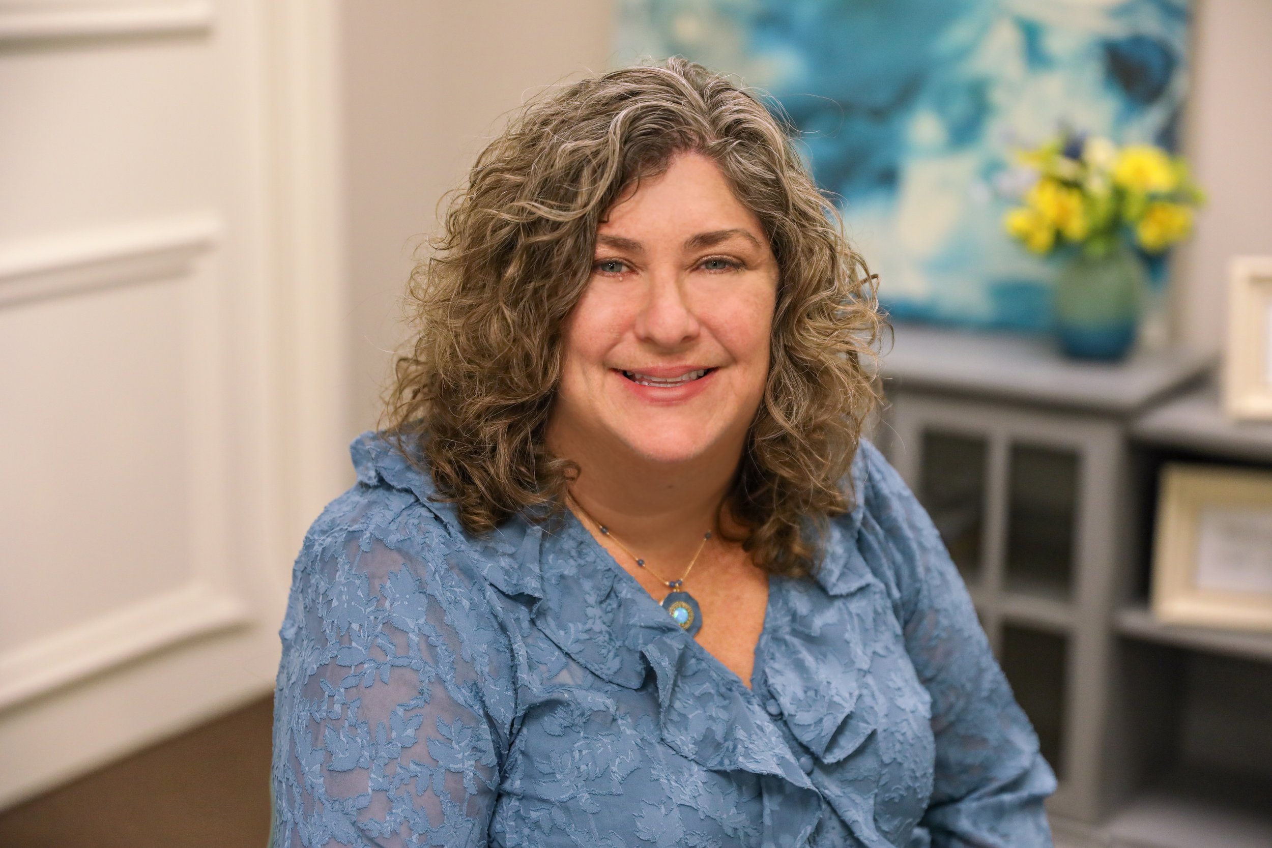 A woman with curly hair wearing a blue lace dress and a necklace, smiling in an indoor setting with a kitchen and yellow flowers in the background.
