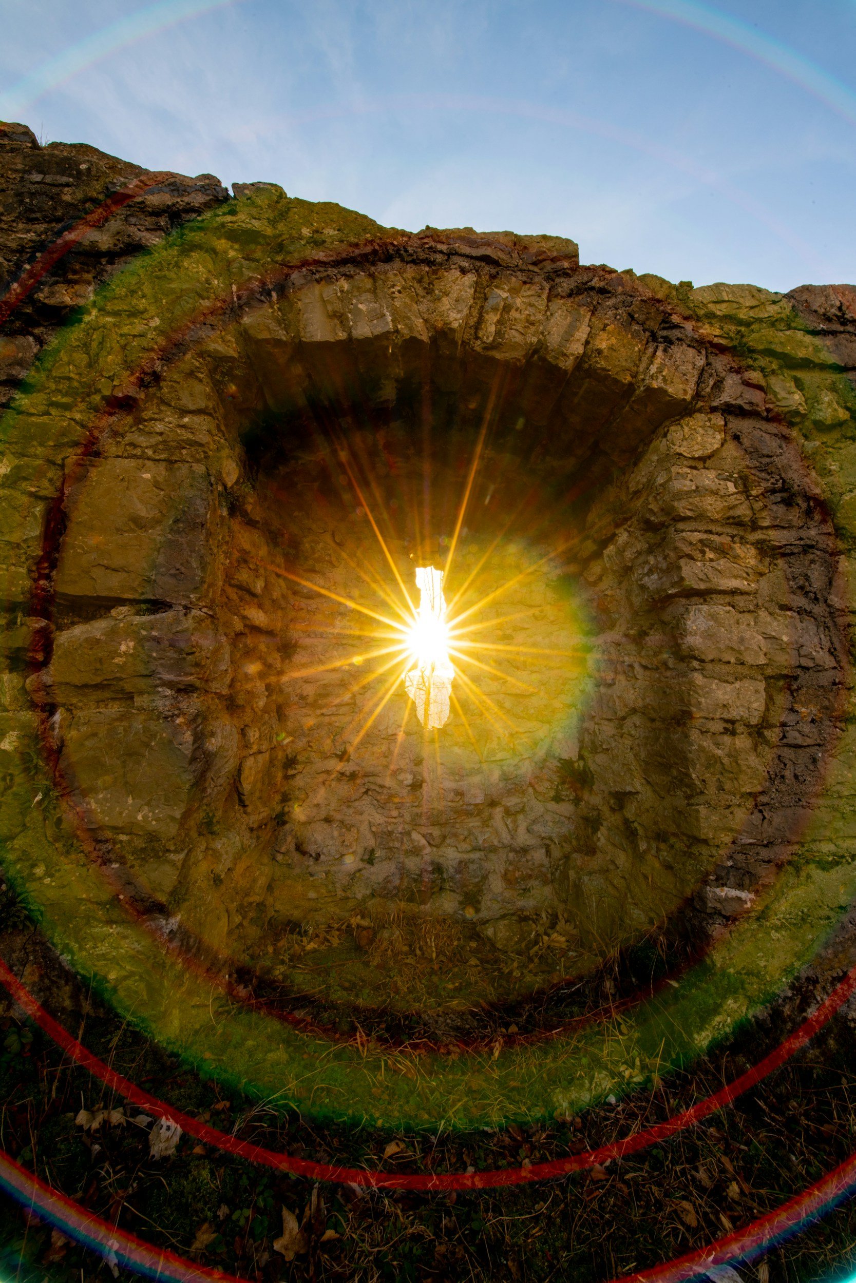 Sunlight shines through an ancient stone archway, creating a starburst effect and rainbow lens flares in a clear blue sky.
