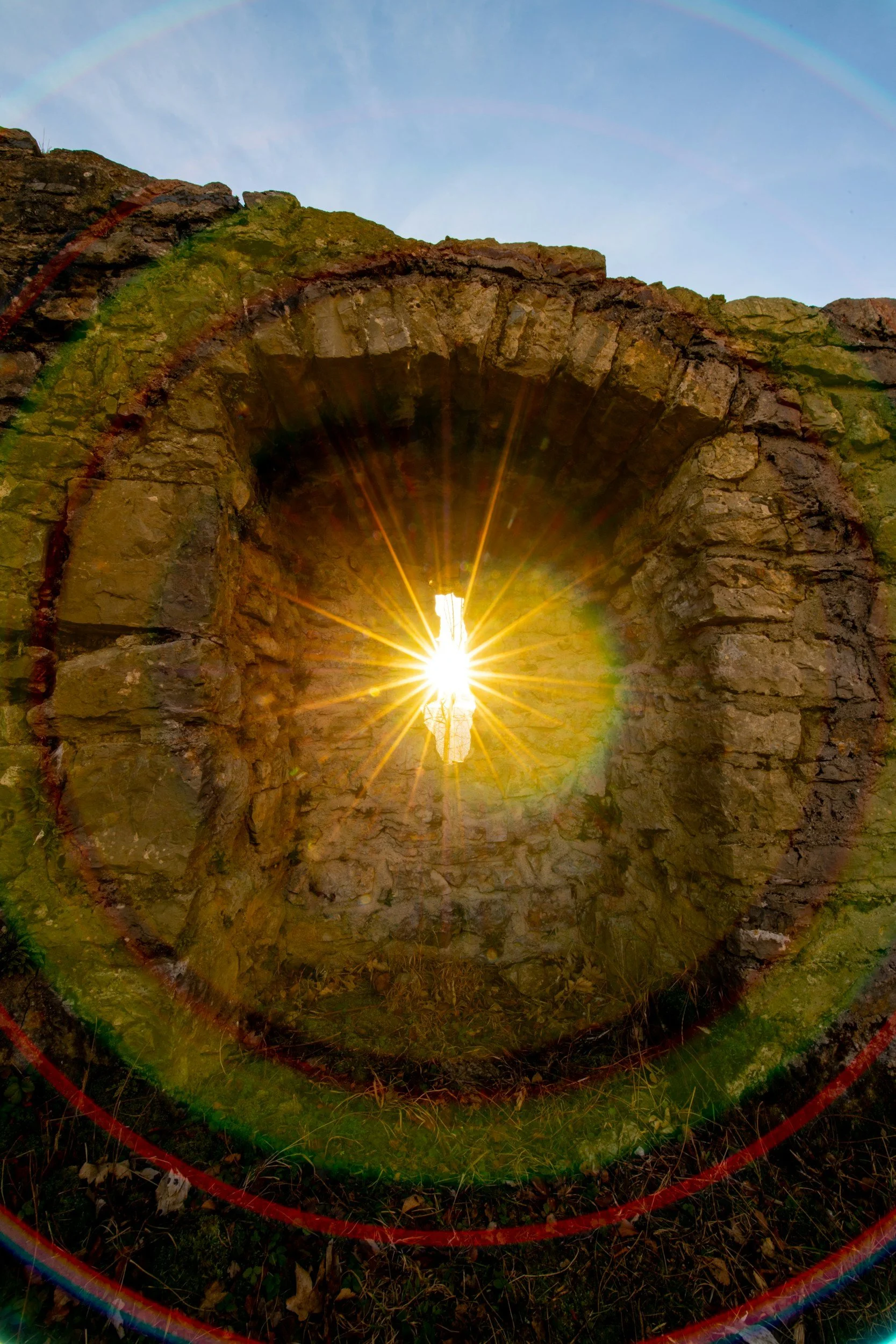 Sun setting through a circular stone window in an ancient stone structure, with a rainbow visible in the sky above.