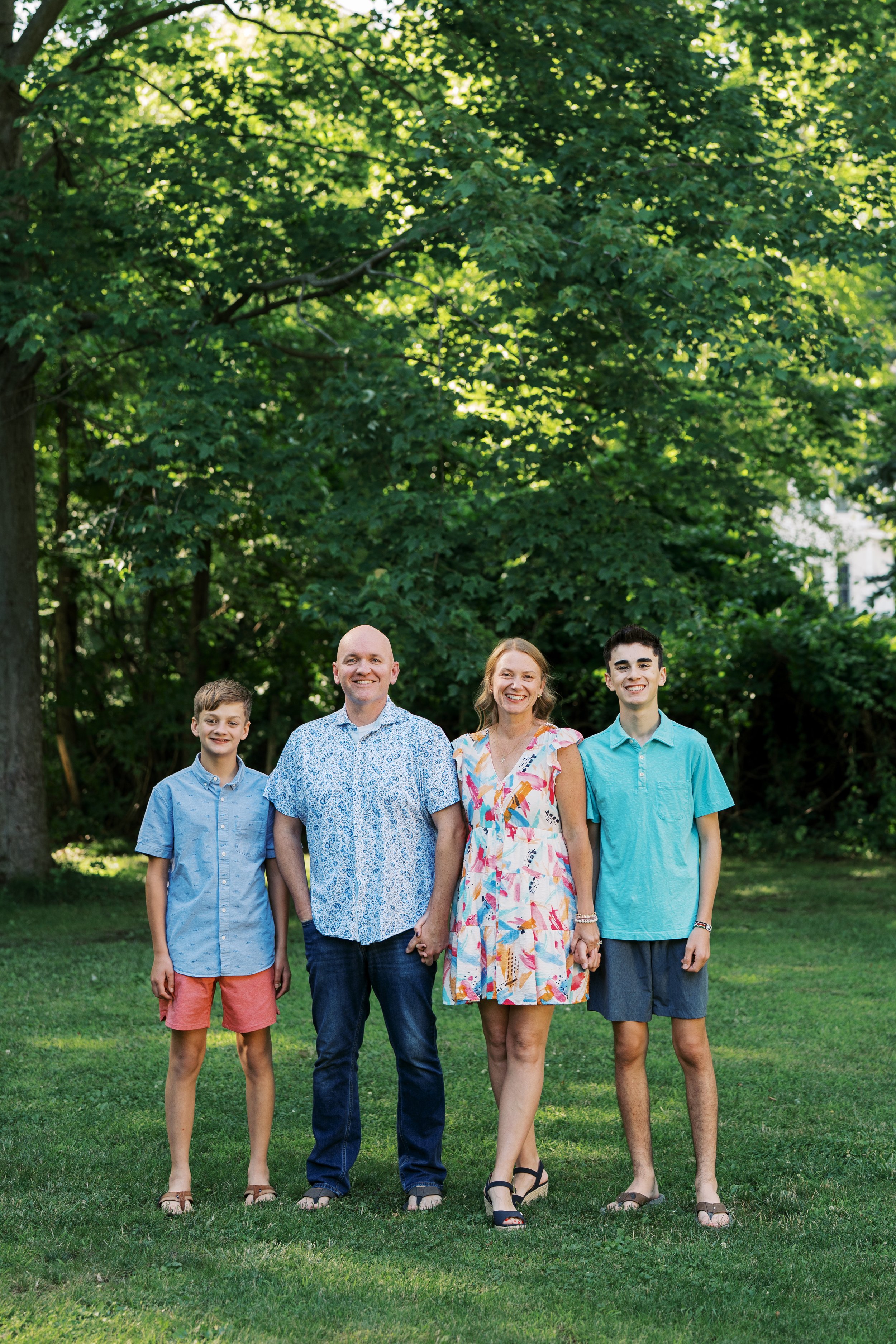 a family of five standing outdoors on a grassy area in front of green trees, smiling at the camera, dressed in casual summer clothes.