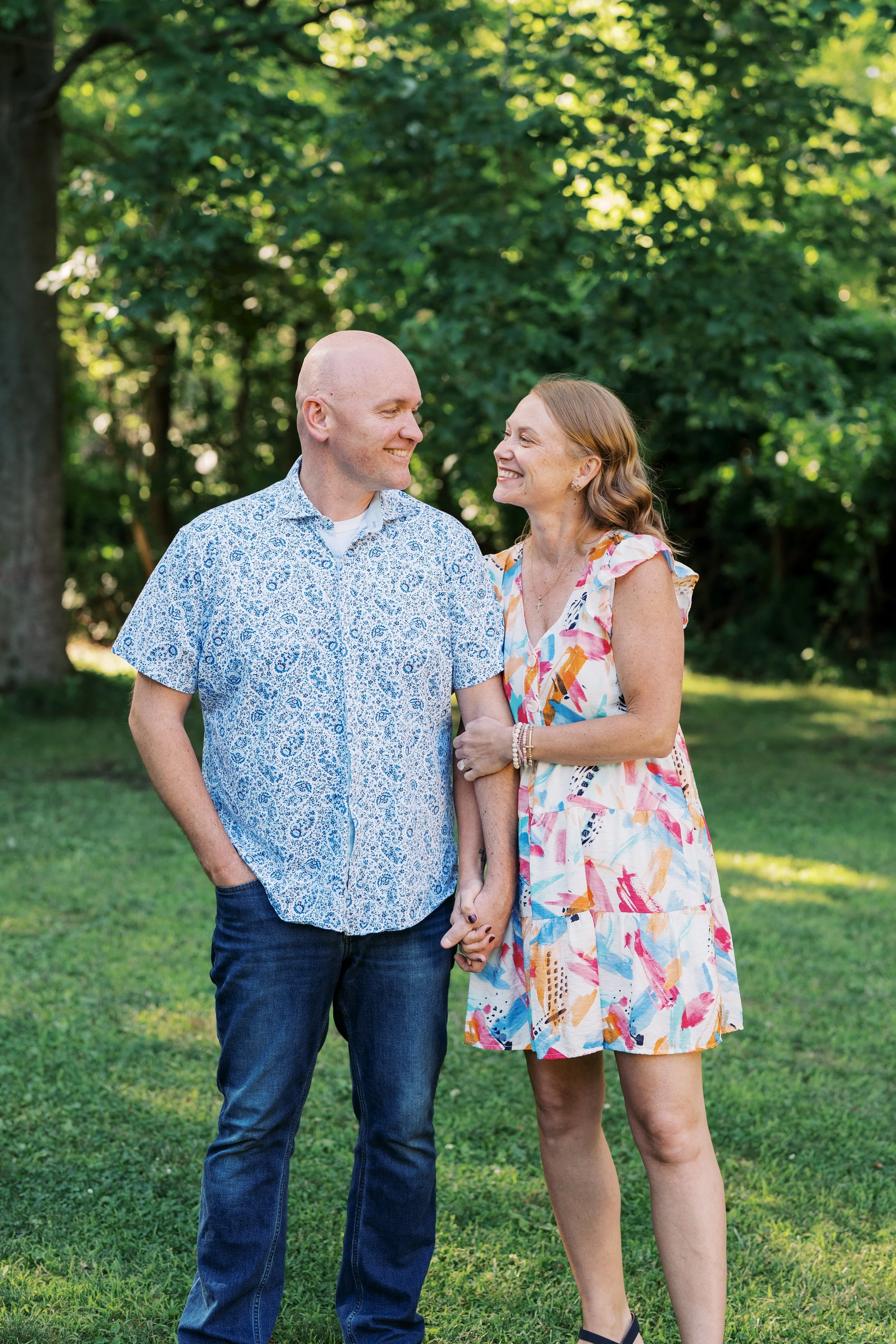 A couple holding hands and smiling at each other outdoors in a park with green trees and grass.