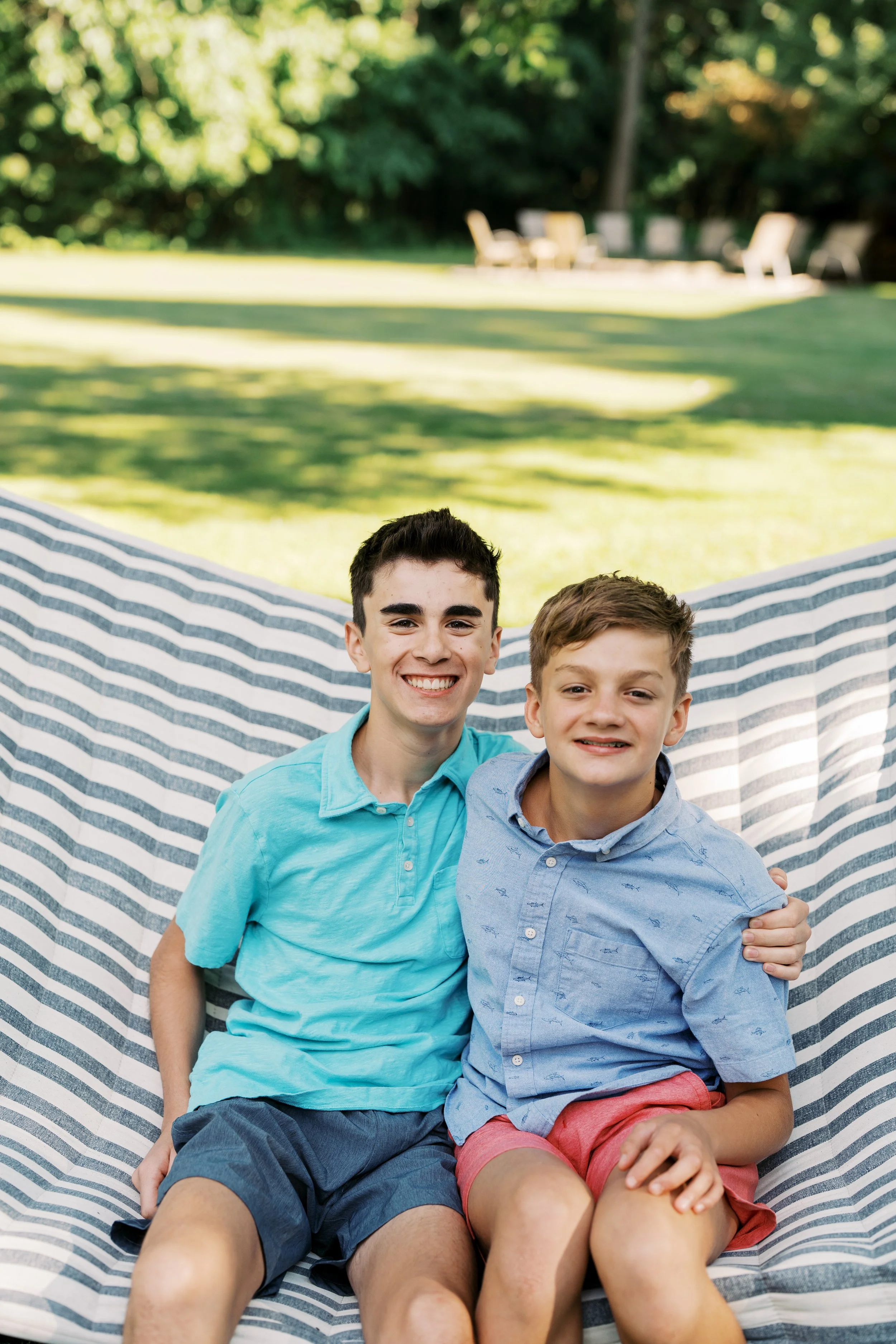 Two young boys smiling and sitting on a striped blanket outside in a grassy area with trees in the background.