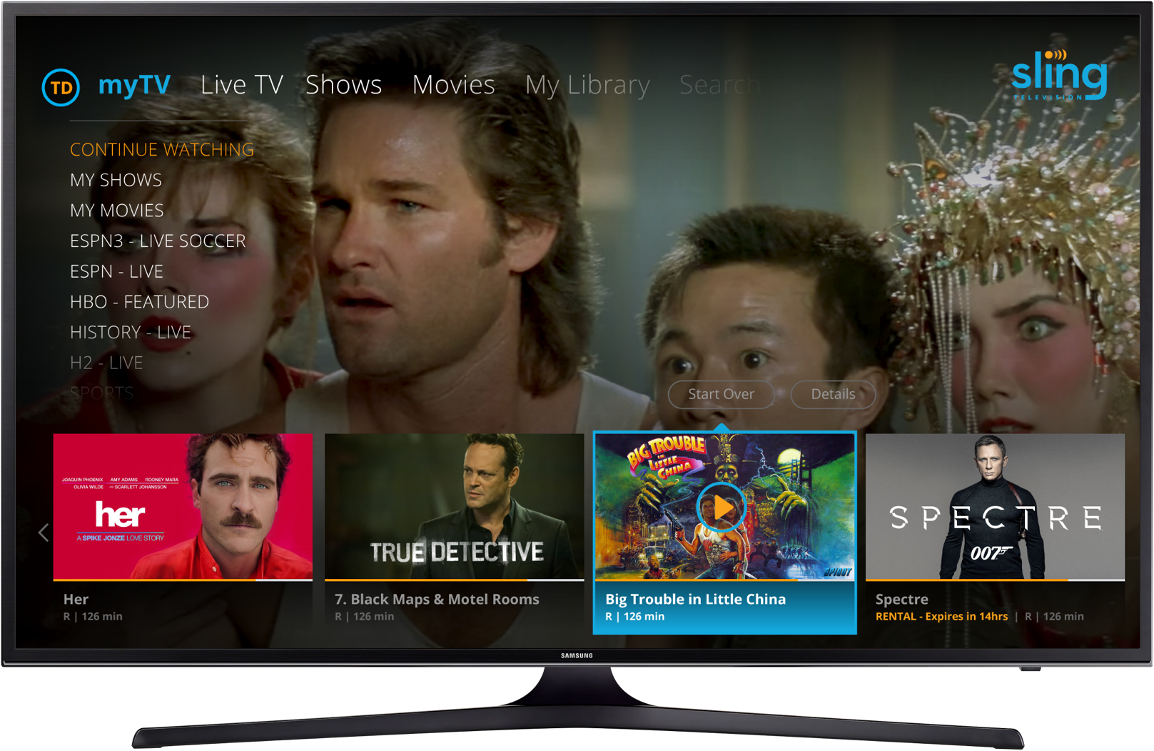 Samsung TV screen displaying a streaming platform interface with movie options, such as 'Her', 'True Detective', 'Big Trouble in Little China', and 'Spectre'. The background features a scene from a movie with three characters, one of whom is wearing a traditional headdress. The menu includes options for live TV, shows, movies, library, and search.