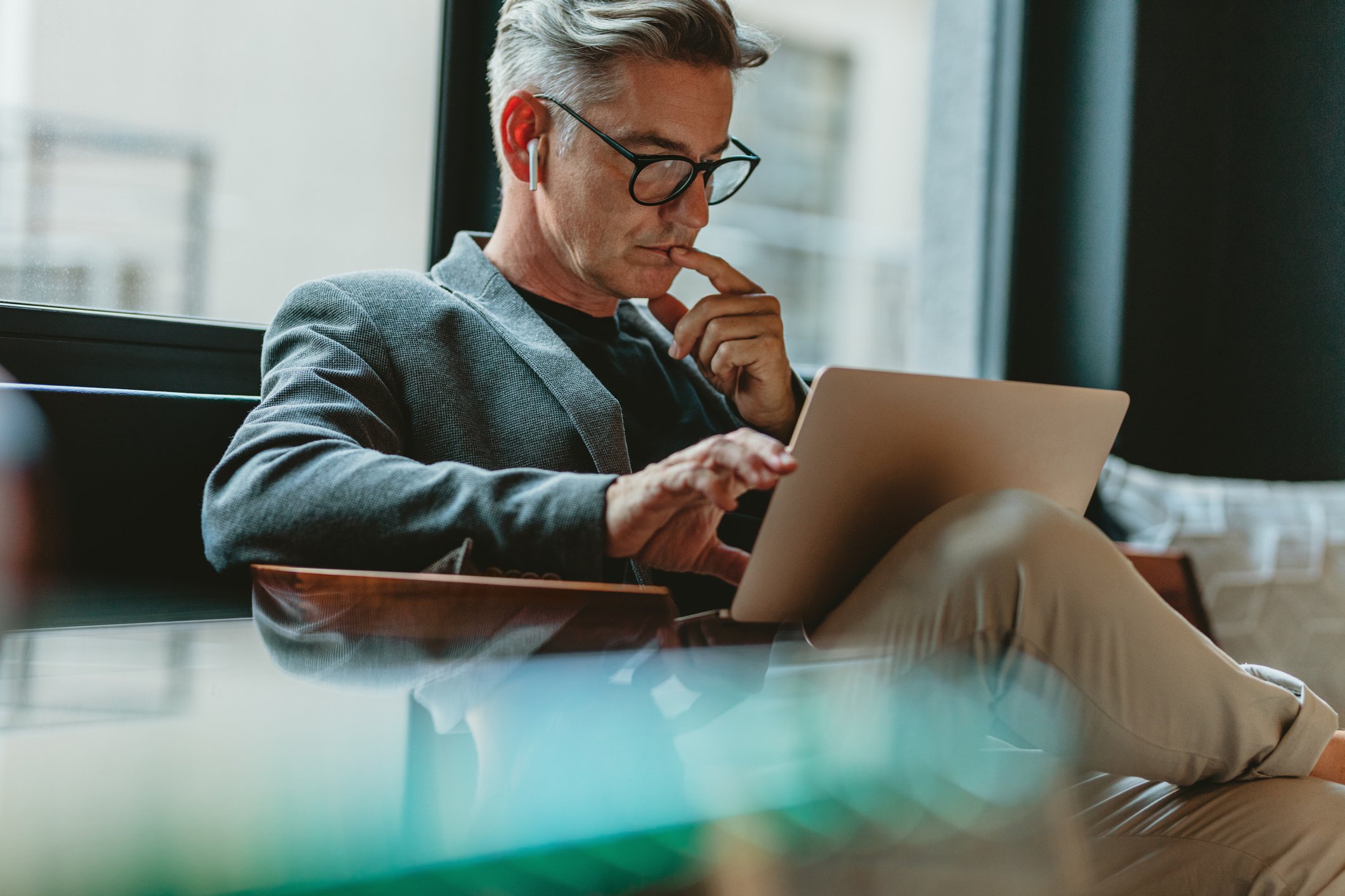 A middle-aged man with gray hair, glasses, and wireless earbuds is sitting on a bench near a large window, looking at a tablet device with a thoughtful expression.
