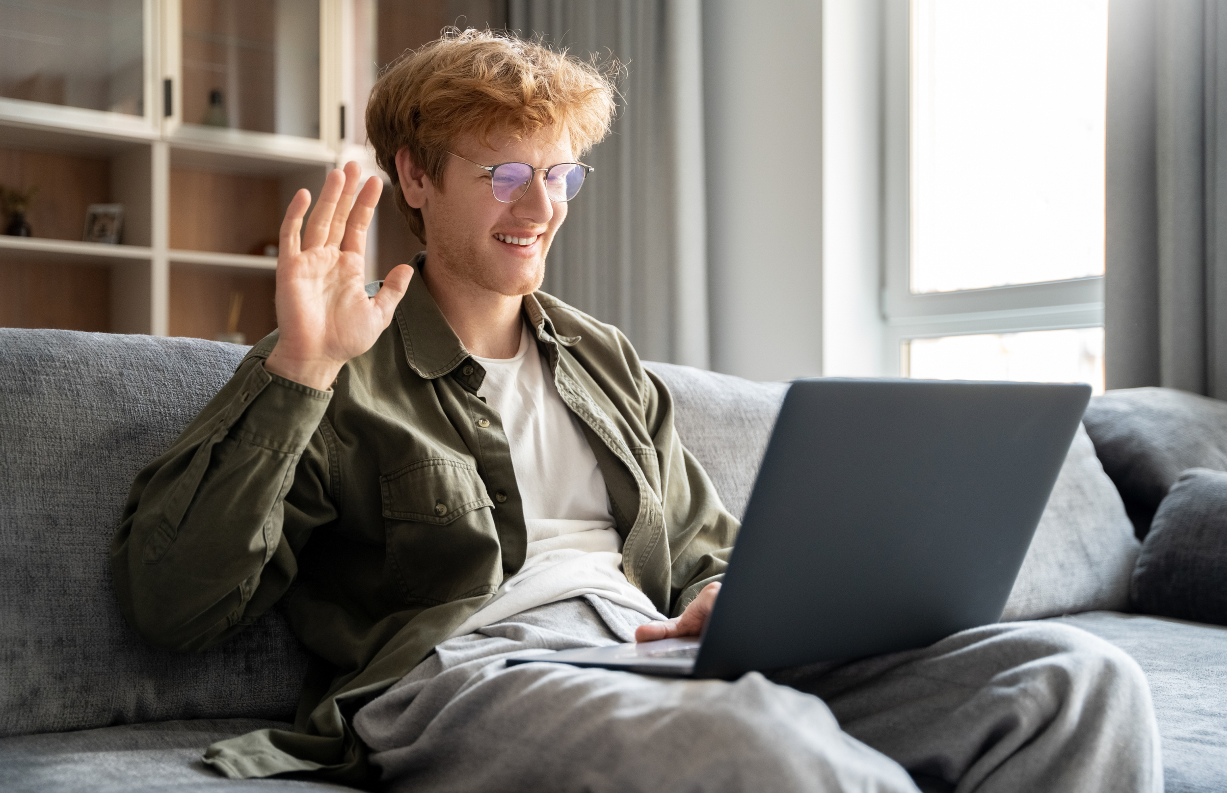 A young man with red hair and glasses sitting on a grey couch, smiling and waving at his laptop screen, in a bright room with a window and bookshelf in the background.